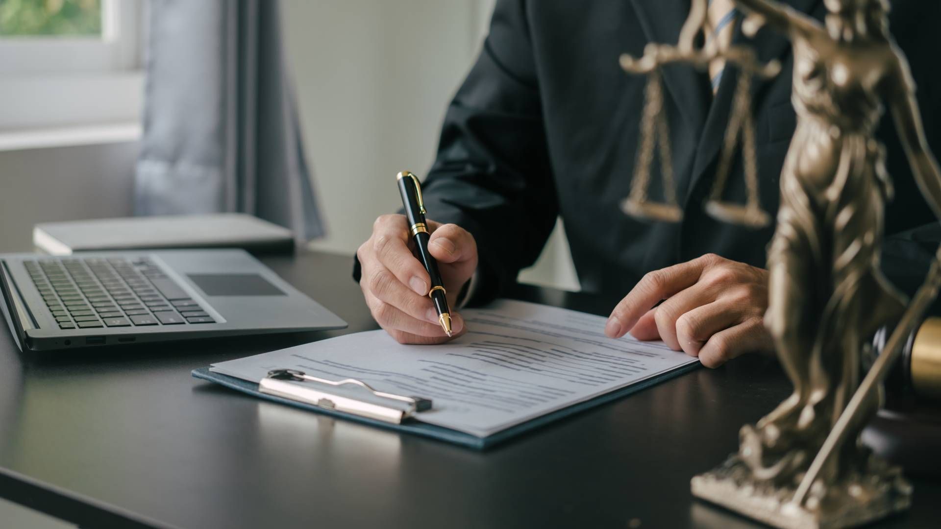 A lawyer is sitting at a desk writing on a clipboard next to a statue of justice.