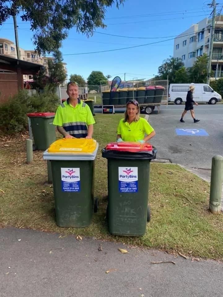 A Man And A Woman Are Standing Next To Garbage Cans In A Parking Lot — Party Bins Pty Ltd In Landsborough, QLD