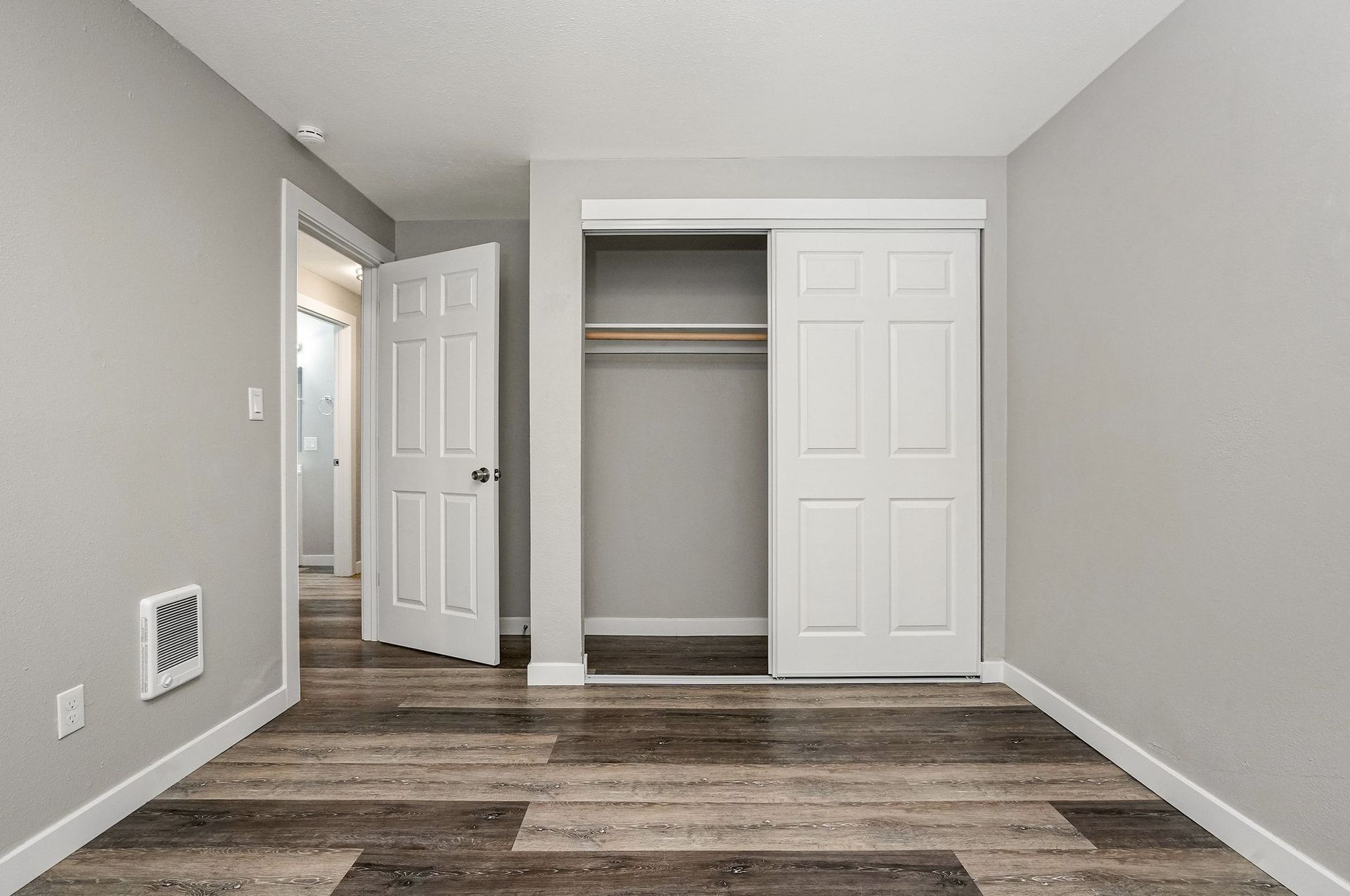 Empty gray bedroom with wood-look flooring, open closet, and open door near a hallway