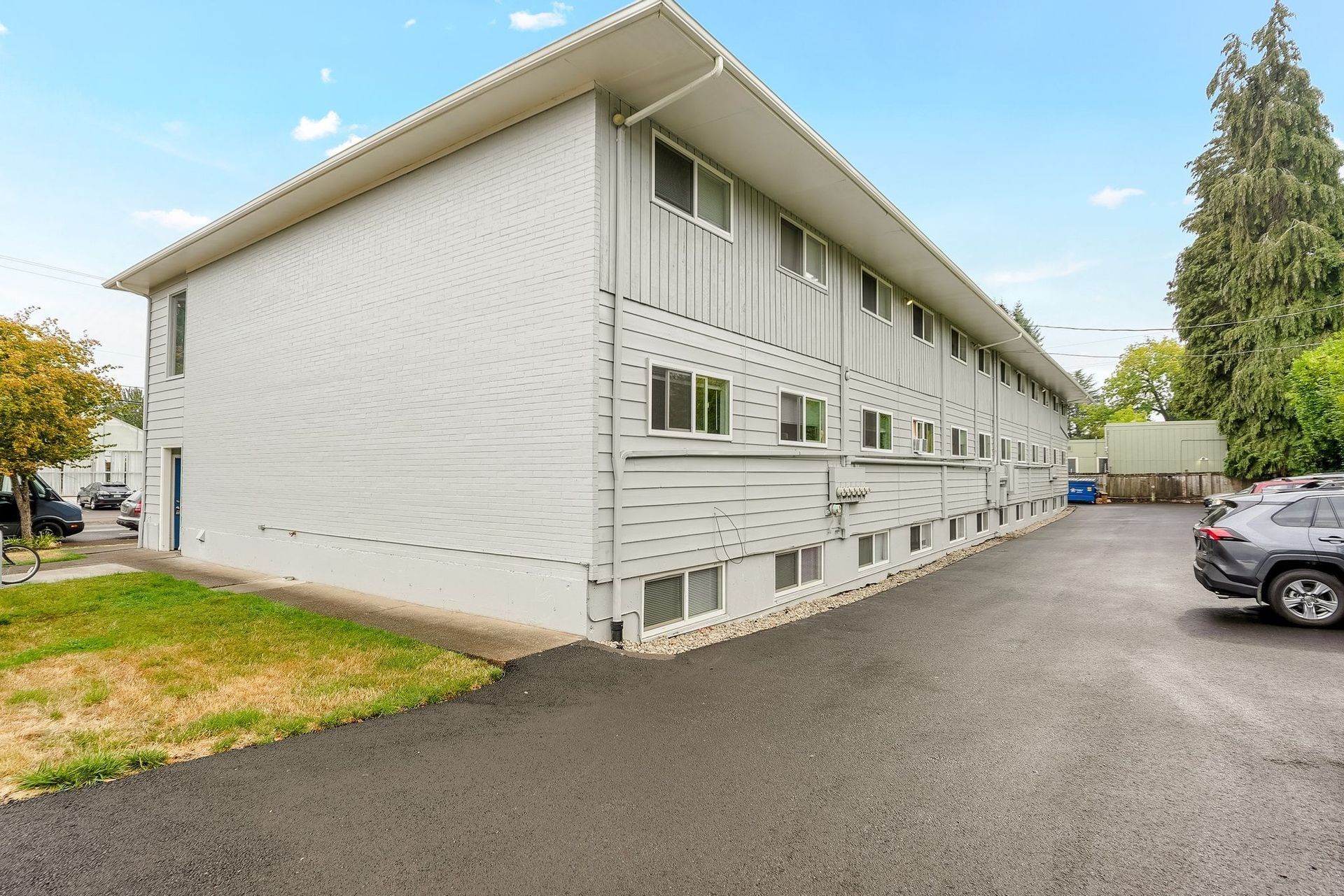 White two-story apartment building with gravel parking lot and parked cars on the side