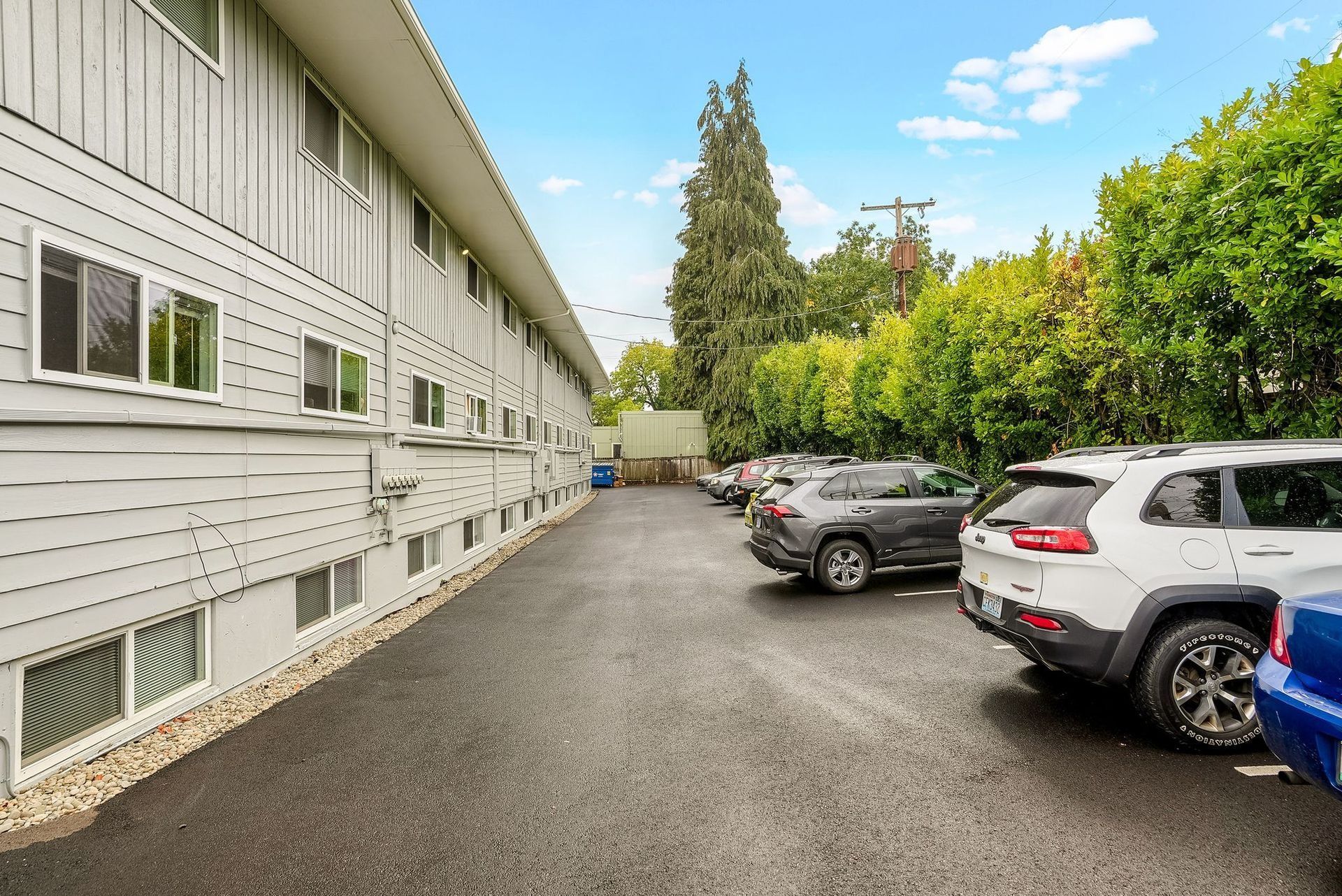 Apartment building parking lot with parked cars along a gravel driveway beside green trees