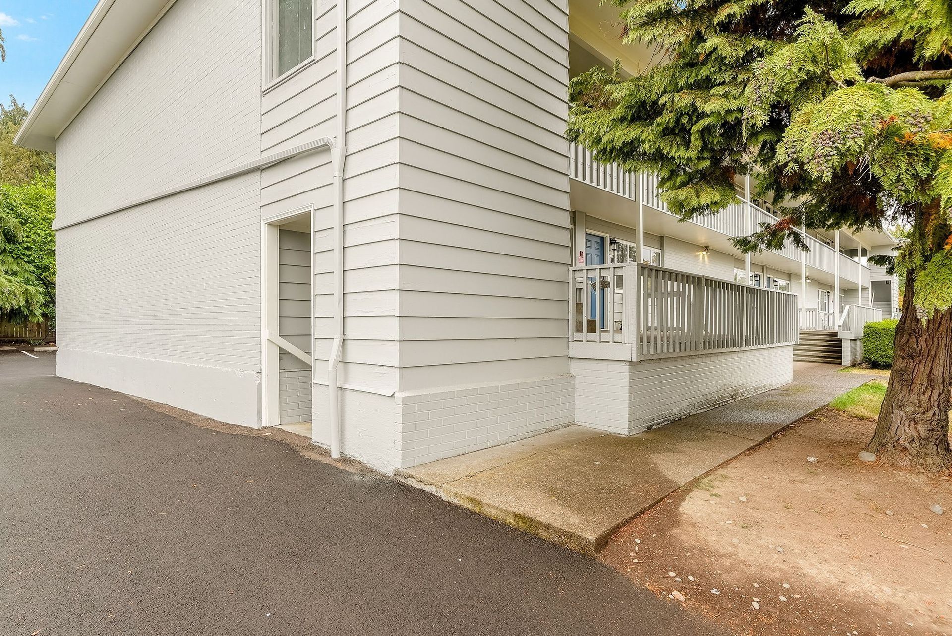 White apartment building with a covered walkway, gravel path, and a large tree beside the entrance.