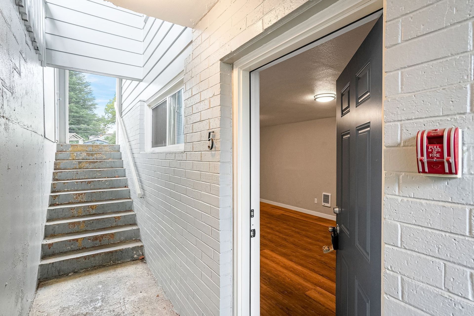 Open apartment entryway with stairs, white brick walls, and a black door partly open to a lit interior.