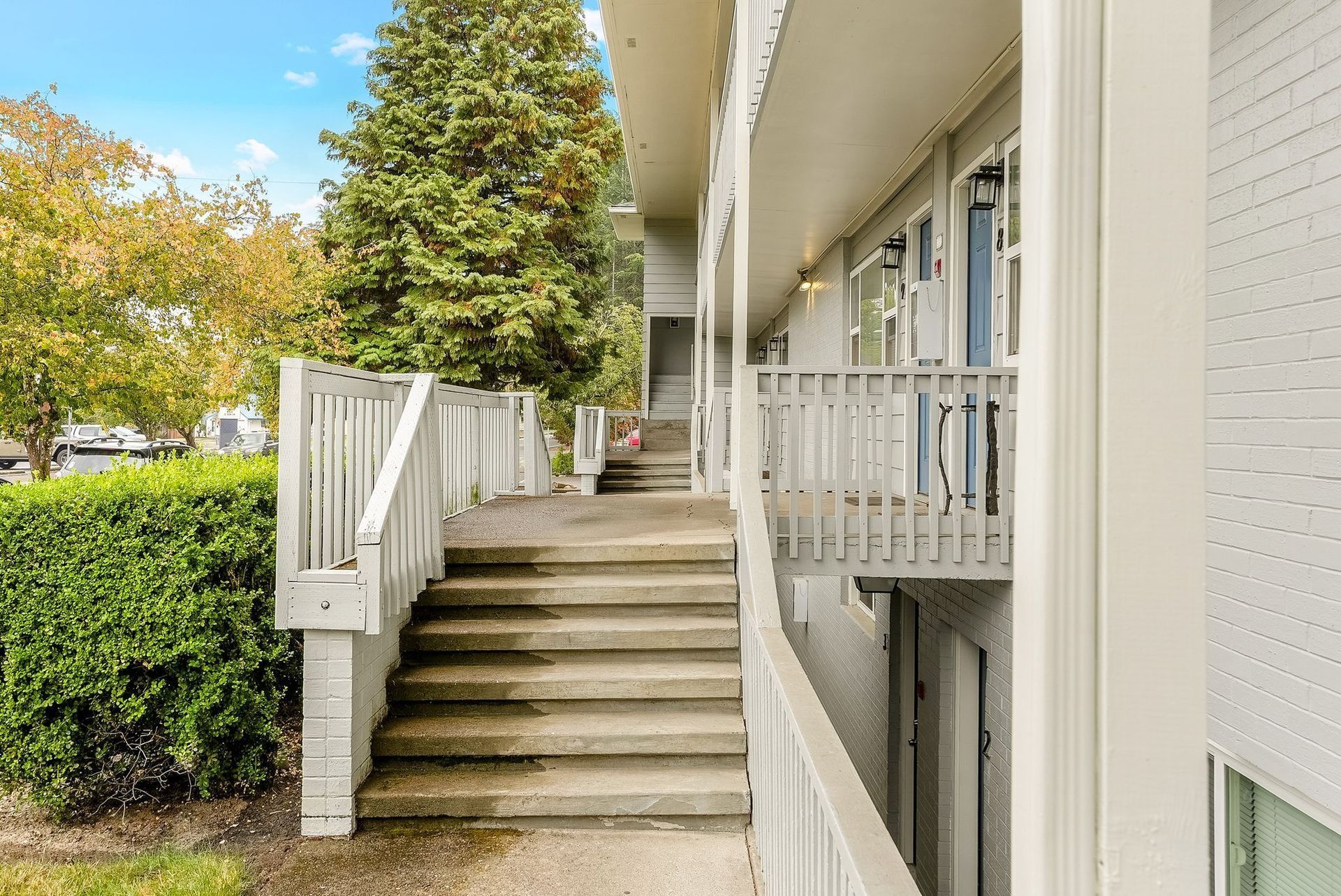 Apartment stairway with white railings beside a walkway and landscaped bushes under trees