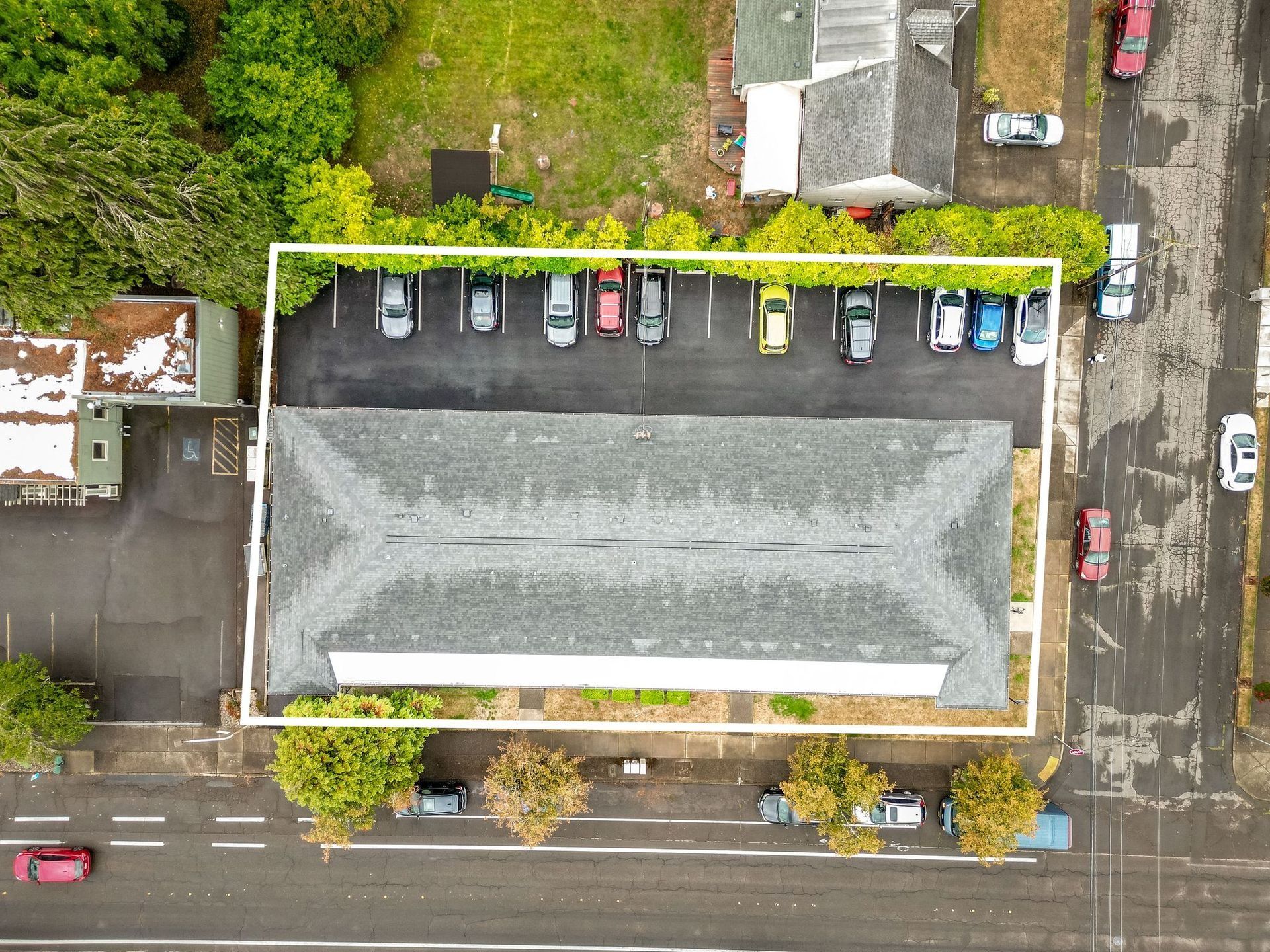 Aerial view of a large building with a parking lot, cars, and trees beside a road.