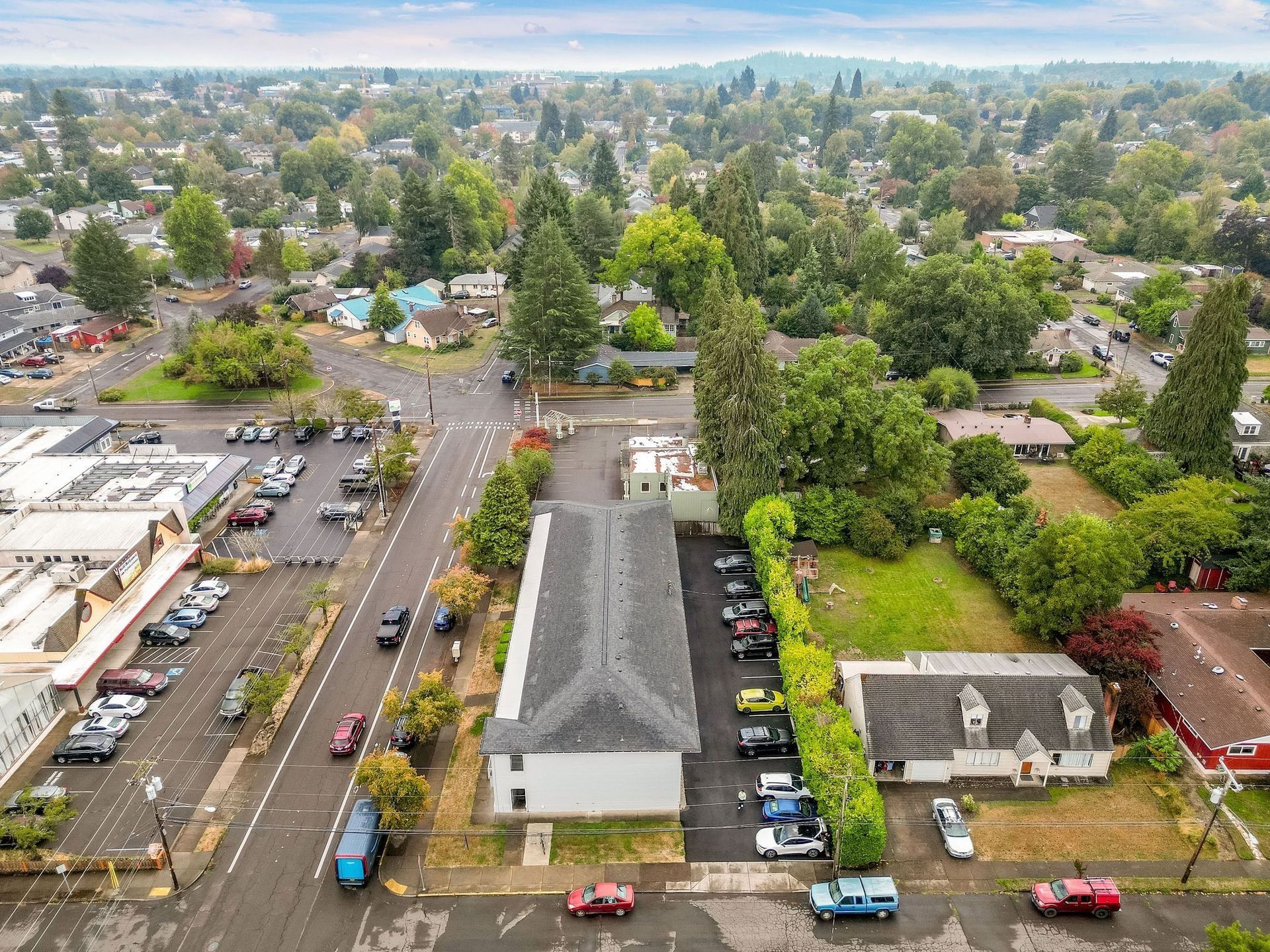 Aerial view of a suburban neighborhood with houses, trees, parked cars, and a cloudy sky.