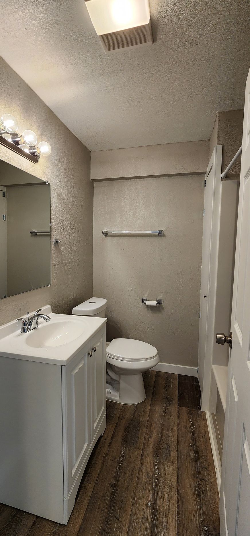 Small bathroom with white vanity, toilet, shower curtain, and wood-look floor under bright ceiling lights