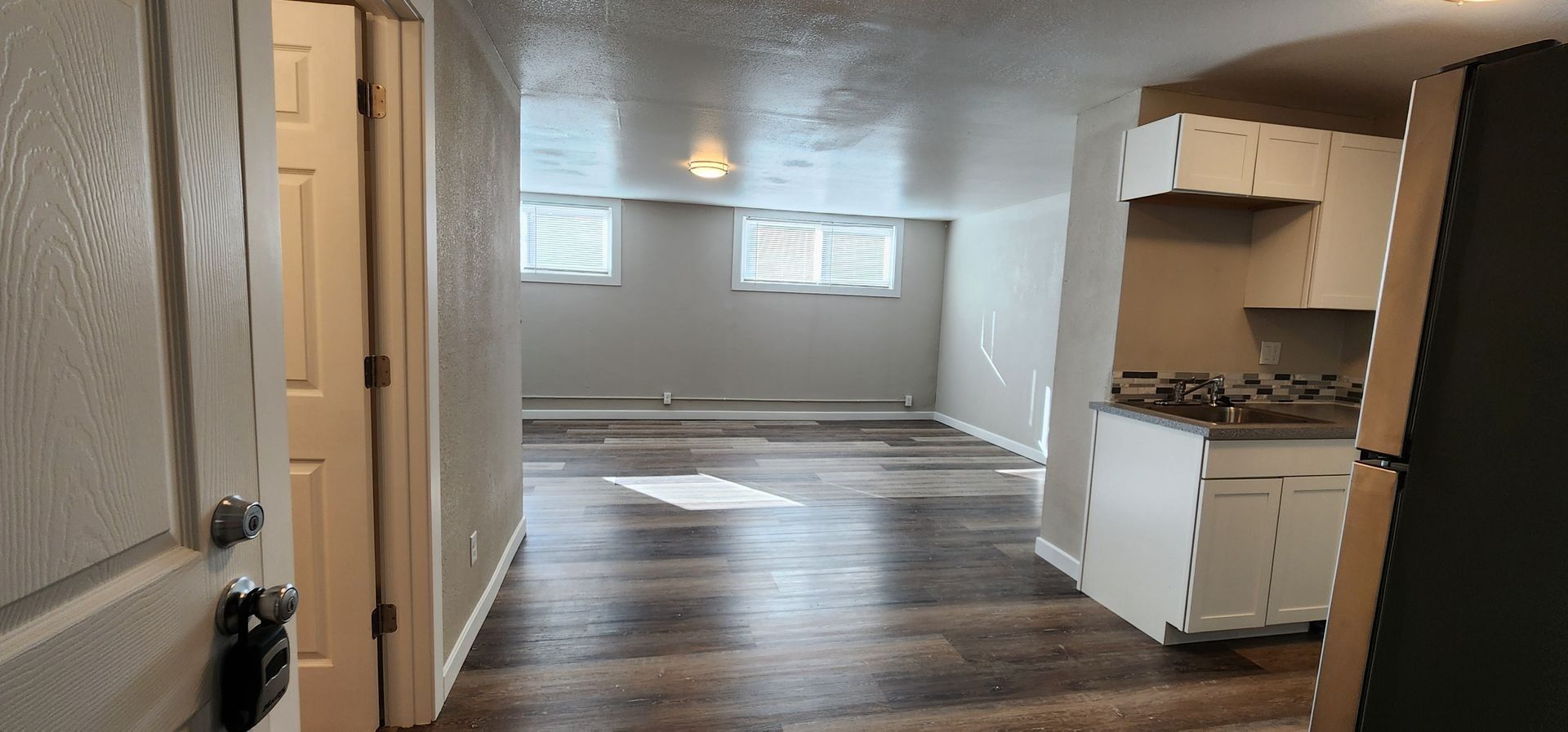 Empty apartment interior with wood floors, small kitchen, and two windows in the back wall