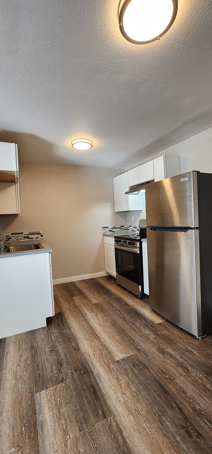 Small modern kitchen with stainless steel fridge, stove, white cabinets, and wood-look flooring.