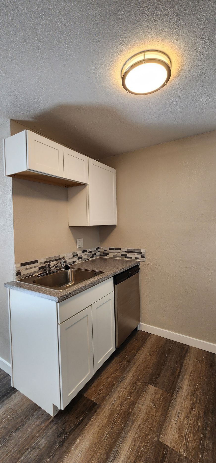 Small empty kitchen with white cabinets, gas stove, beige walls, and wood-look flooring under a ceiling light
