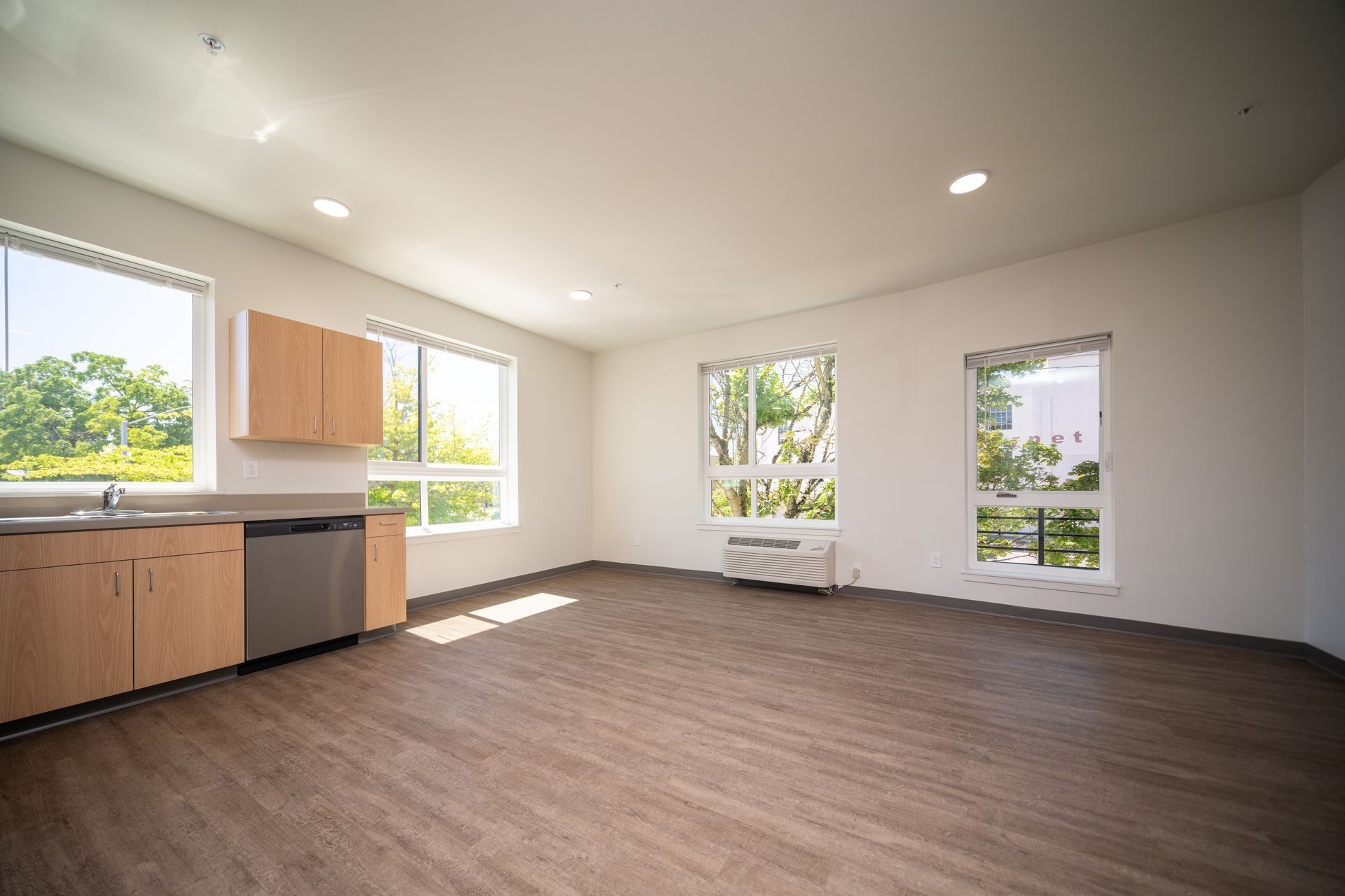 Empty open-plan kitchen and living room with wood cabinets, dark flooring, and large windows.