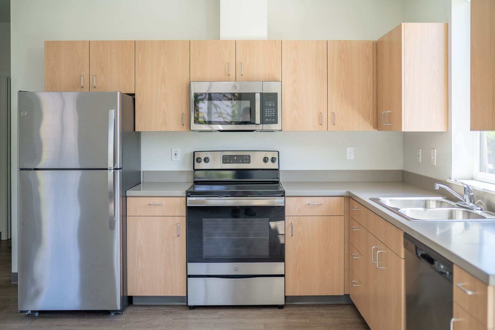 Modern kitchen with stainless steel appliances, light wood cabinets, and a double sink under a window.