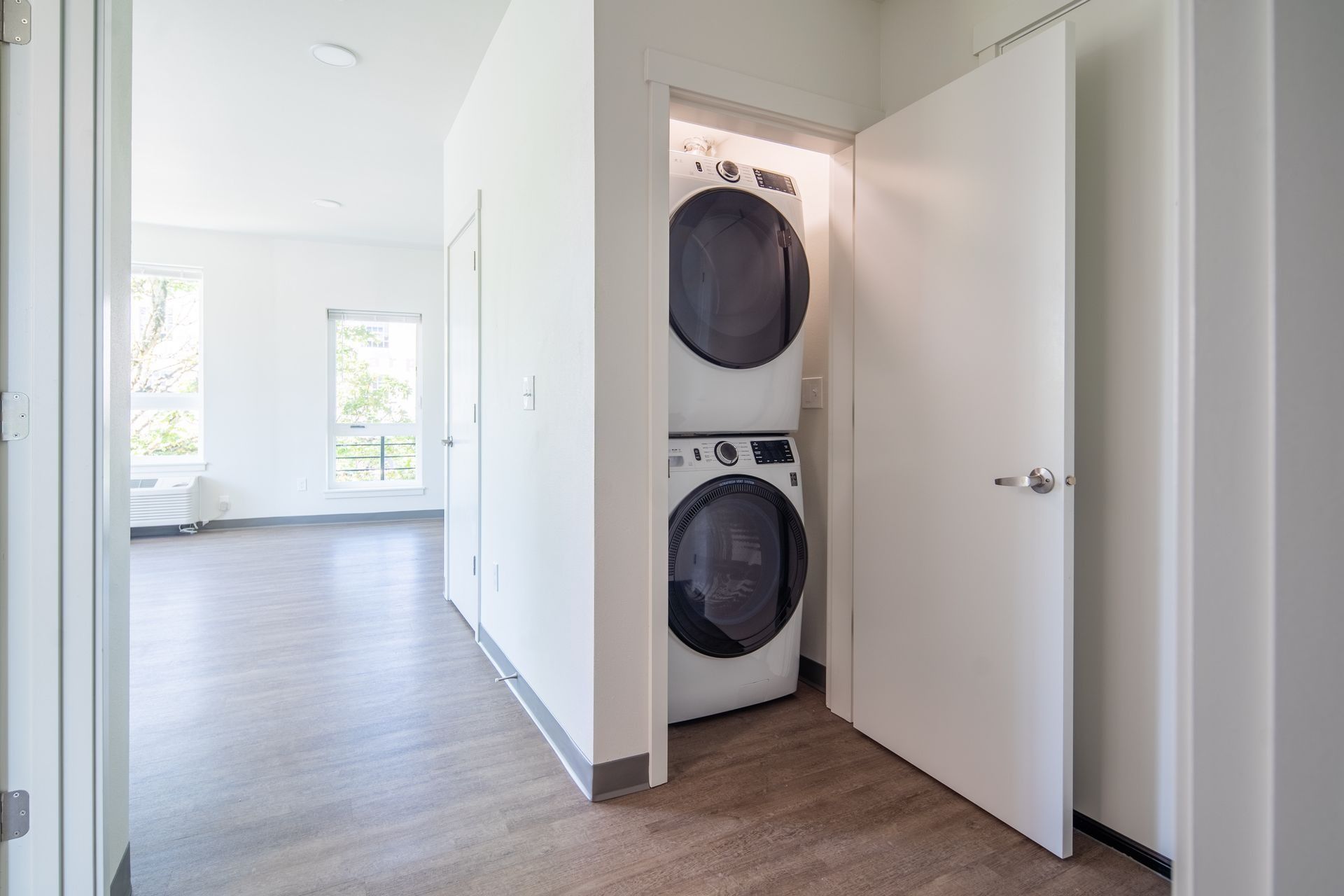 Hallway with stacked washer and dryer in a closet, leading to a bright room with windows.