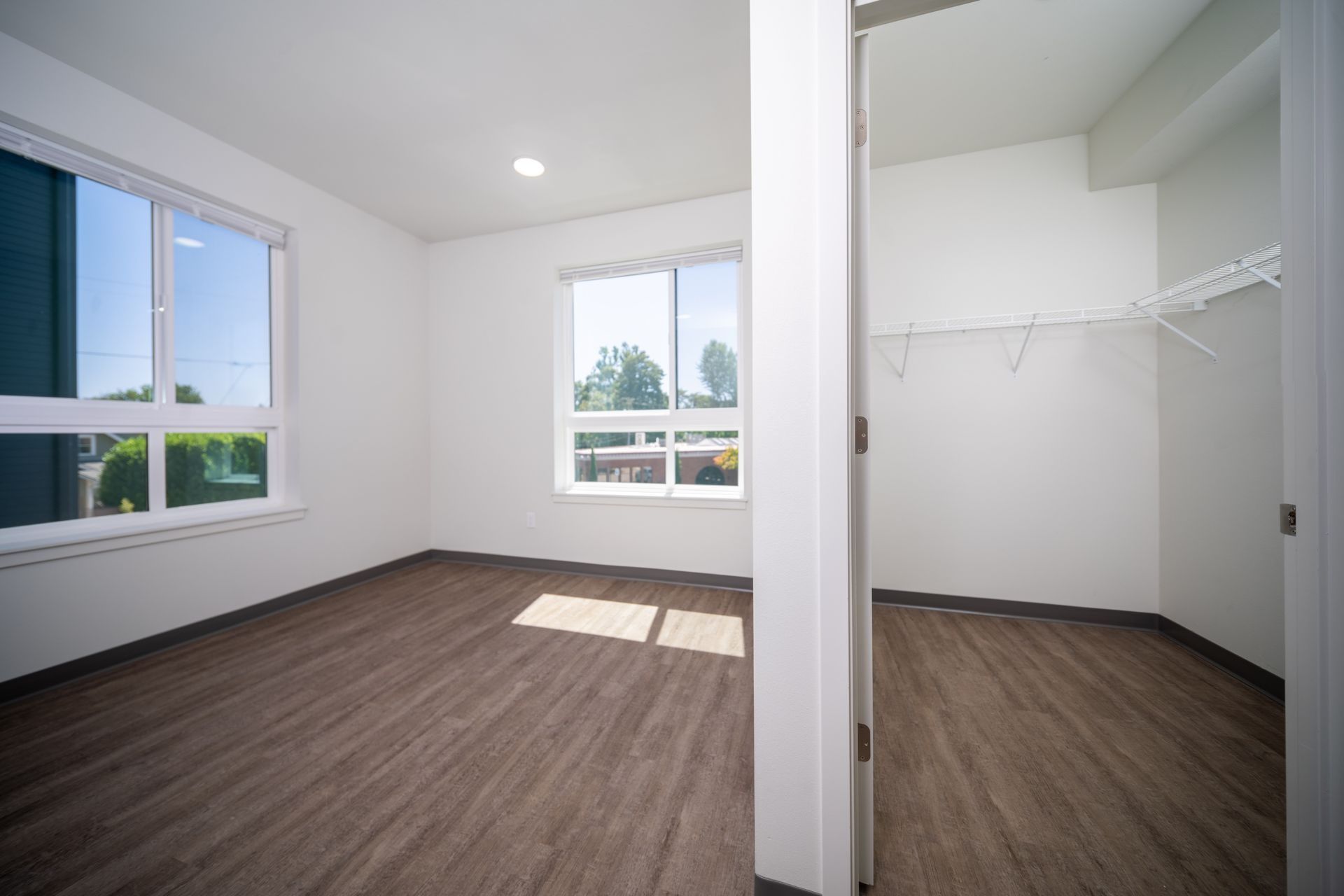 Empty modern bedroom with white walls, wood floors, large windows, and a walk-in closet.