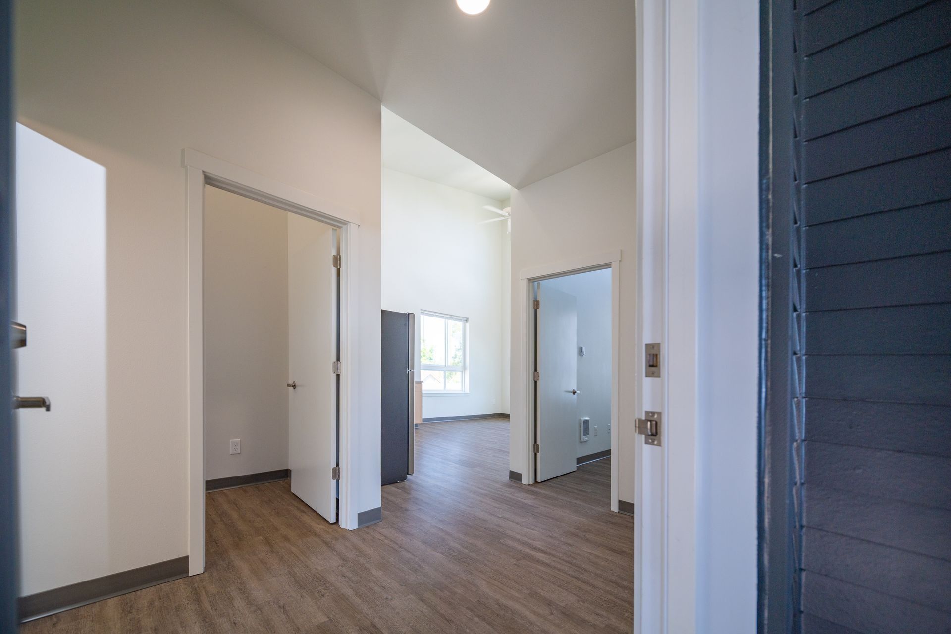 Empty apartment hallway with wood floors, white walls, and open doorways leading to a bright room