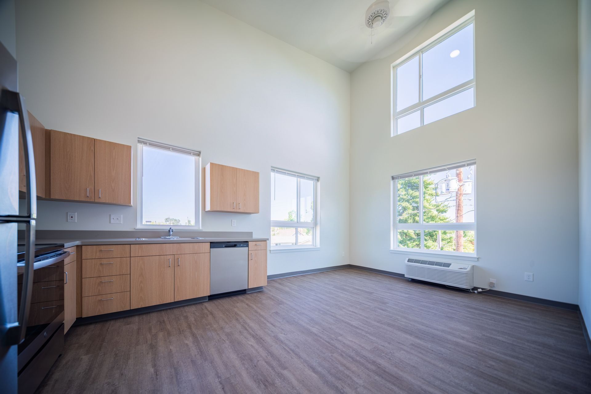 Empty modern kitchen with wood cabinets, dark flooring, high ceiling, and bright windows.