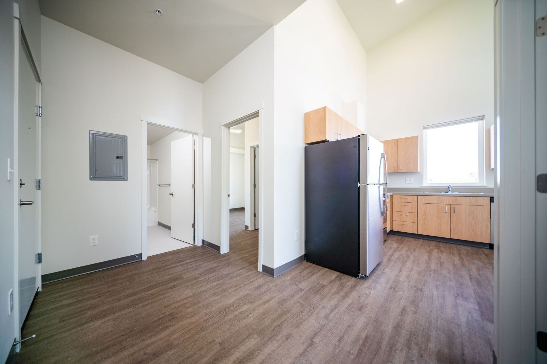 Empty modern apartment room with wood floors, white walls, a kitchen sink, and a black refrigerator