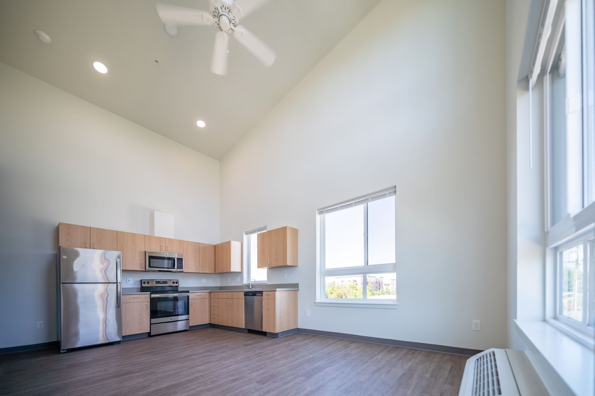 Bright modern kitchen-living space with stainless appliances, wood cabinets, high ceiling, and large windows