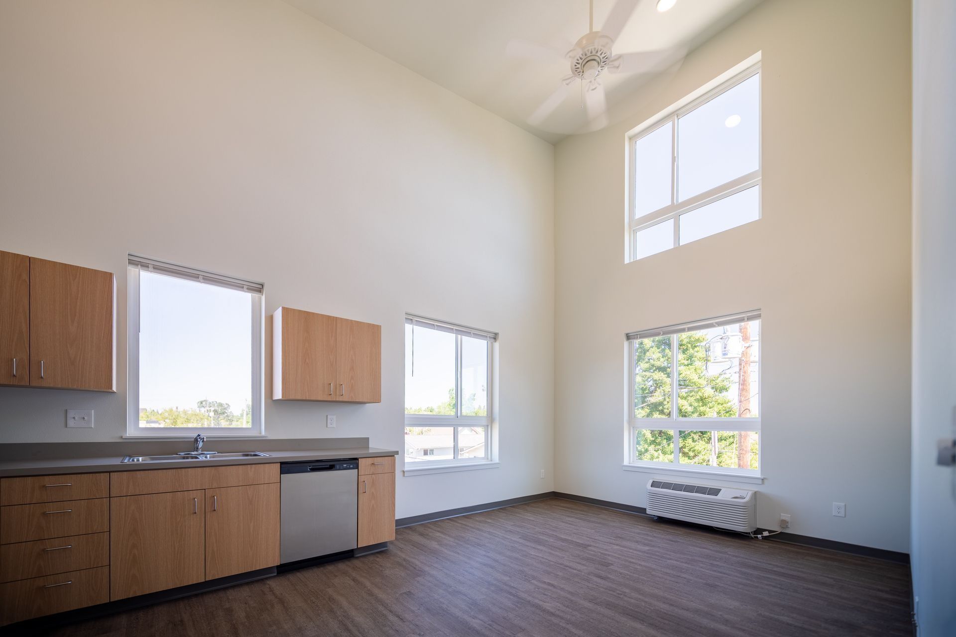 Spacious empty room with kitchen cabinets, large windows, high ceiling, and dark wood floor