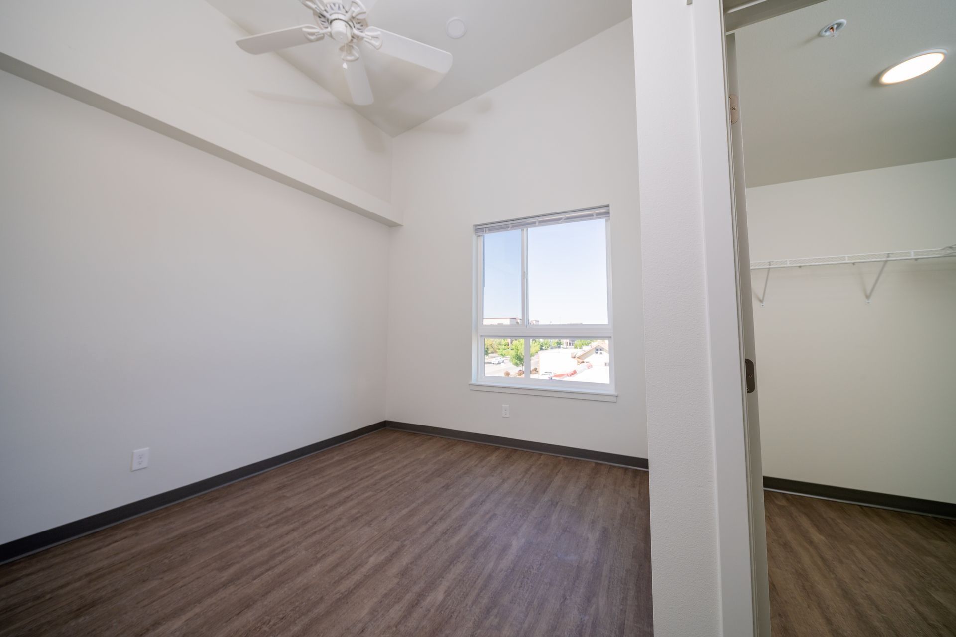 Empty bedroom with white walls, wood-look flooring, ceiling fan, and a window with a closet nook.