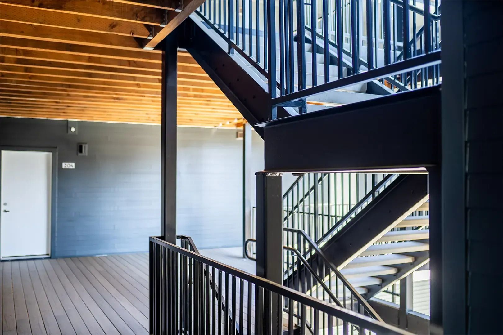 Interior stairwell with black railings, wooden ceiling beams, and a gray door on a balcony.
