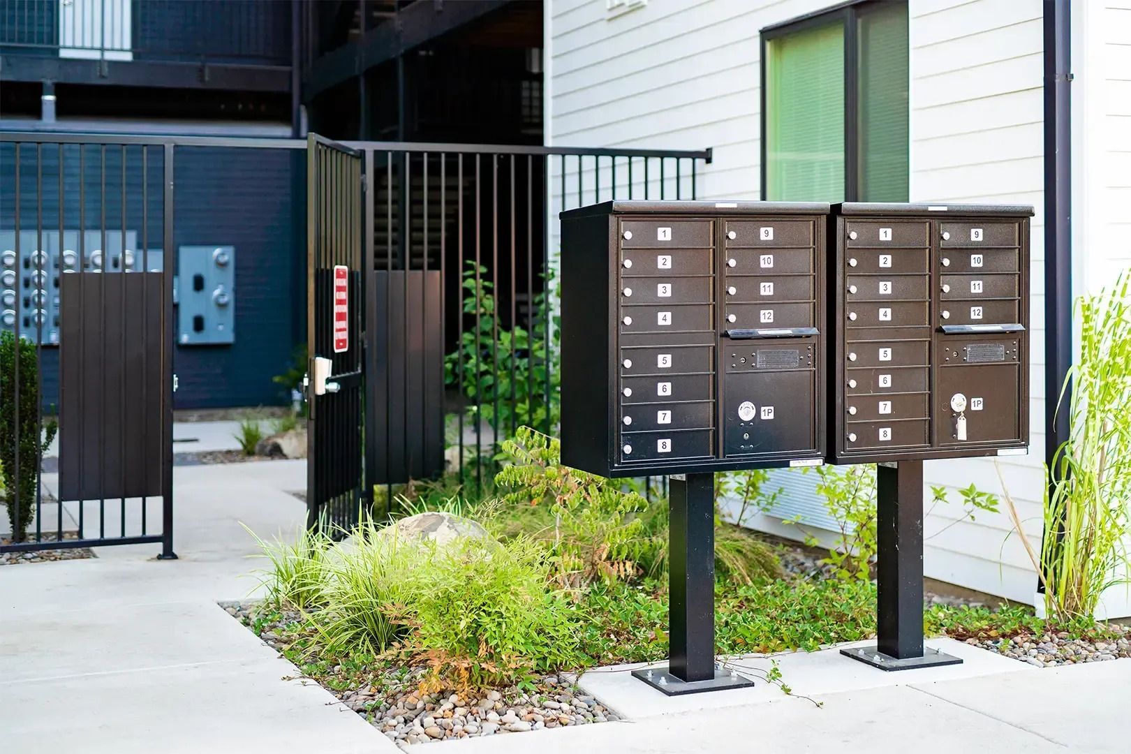 Brown apartment mailboxes beside a gated building entrance with a walkway and plants