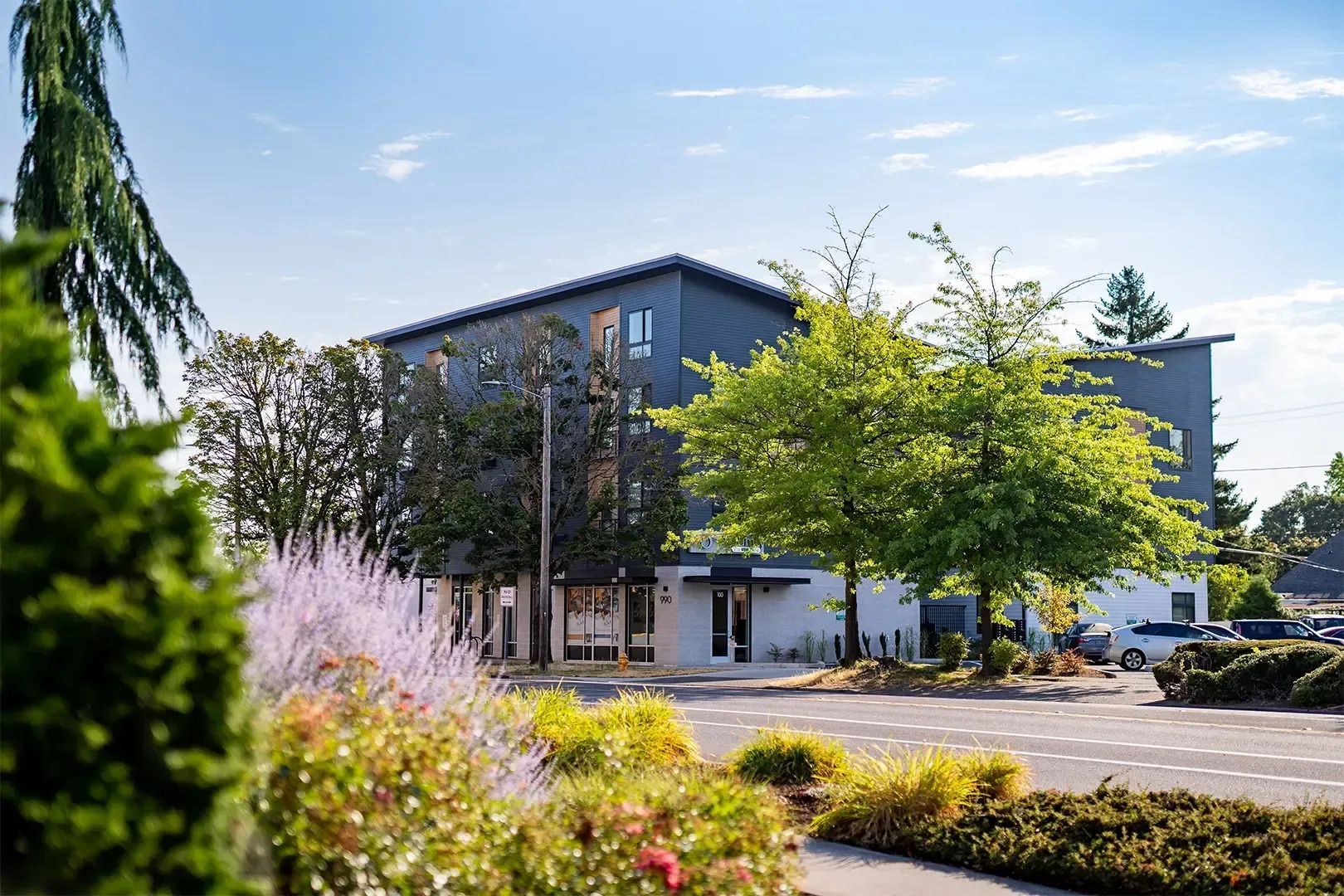 Modern office building with trees, flowering plants, and parked cars on a sunny day