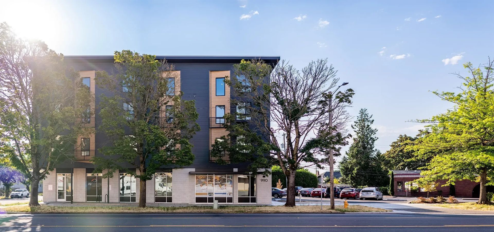 Modern apartment building with trees, storefronts, and street parking on a sunny day