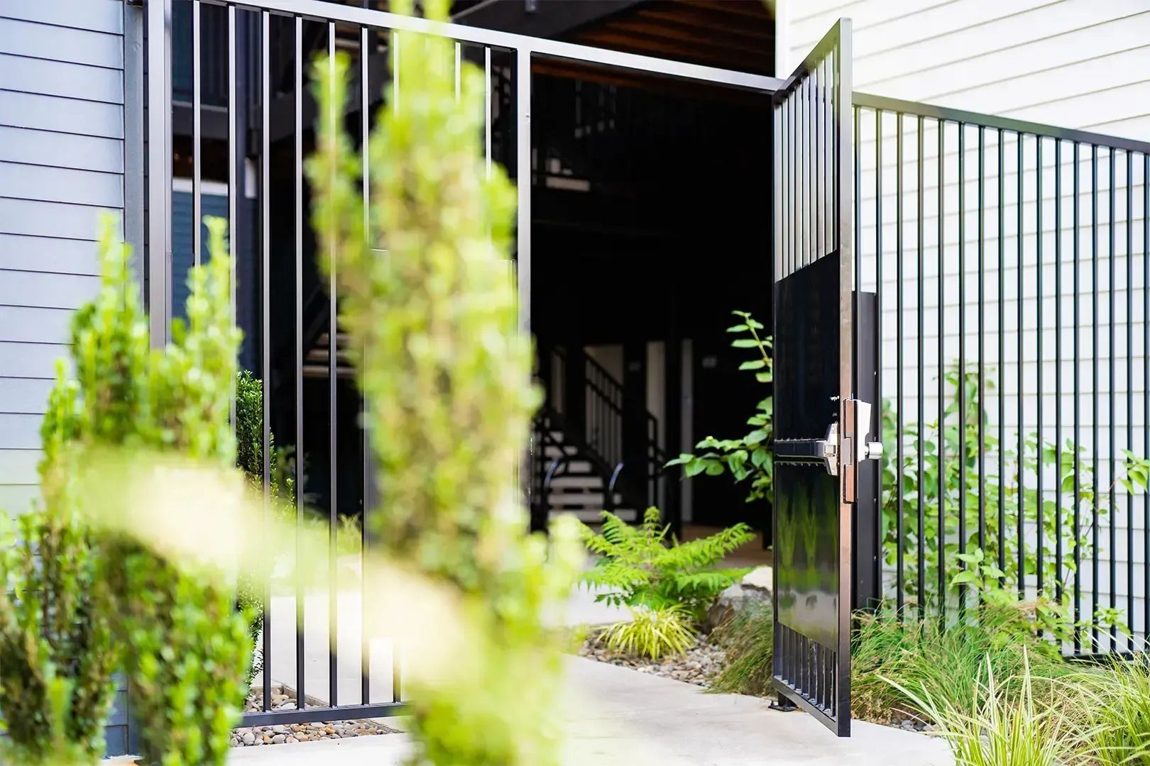 Open black gate beside modern house with narrow garden path and tall plants in front