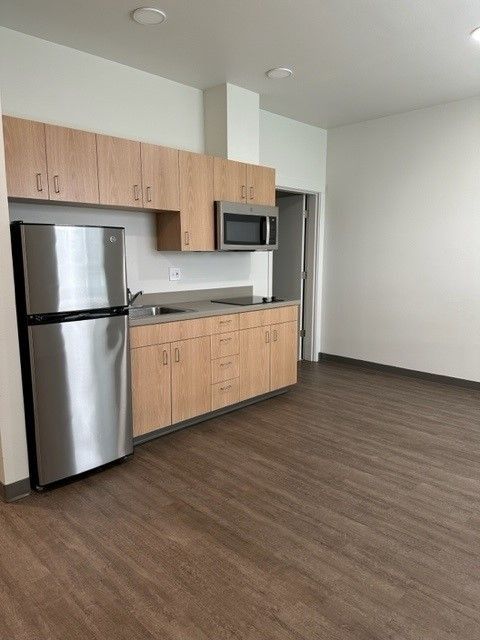 Empty modern apartment kitchen with stainless steel fridge, wood cabinets, and dark flooring