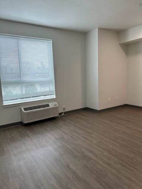 Empty room with gray wood floors, beige walls, and a window air conditioner beneath a window.