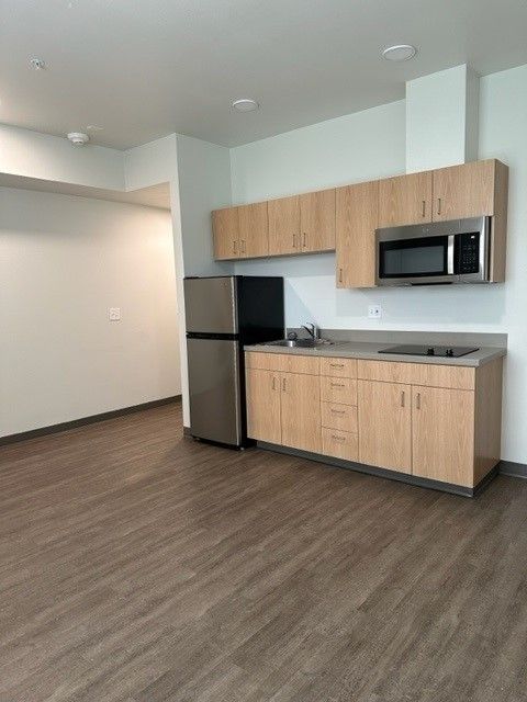 Small modern studio kitchenette with wood cabinets, stainless fridge, microwave, and sink in an empty room