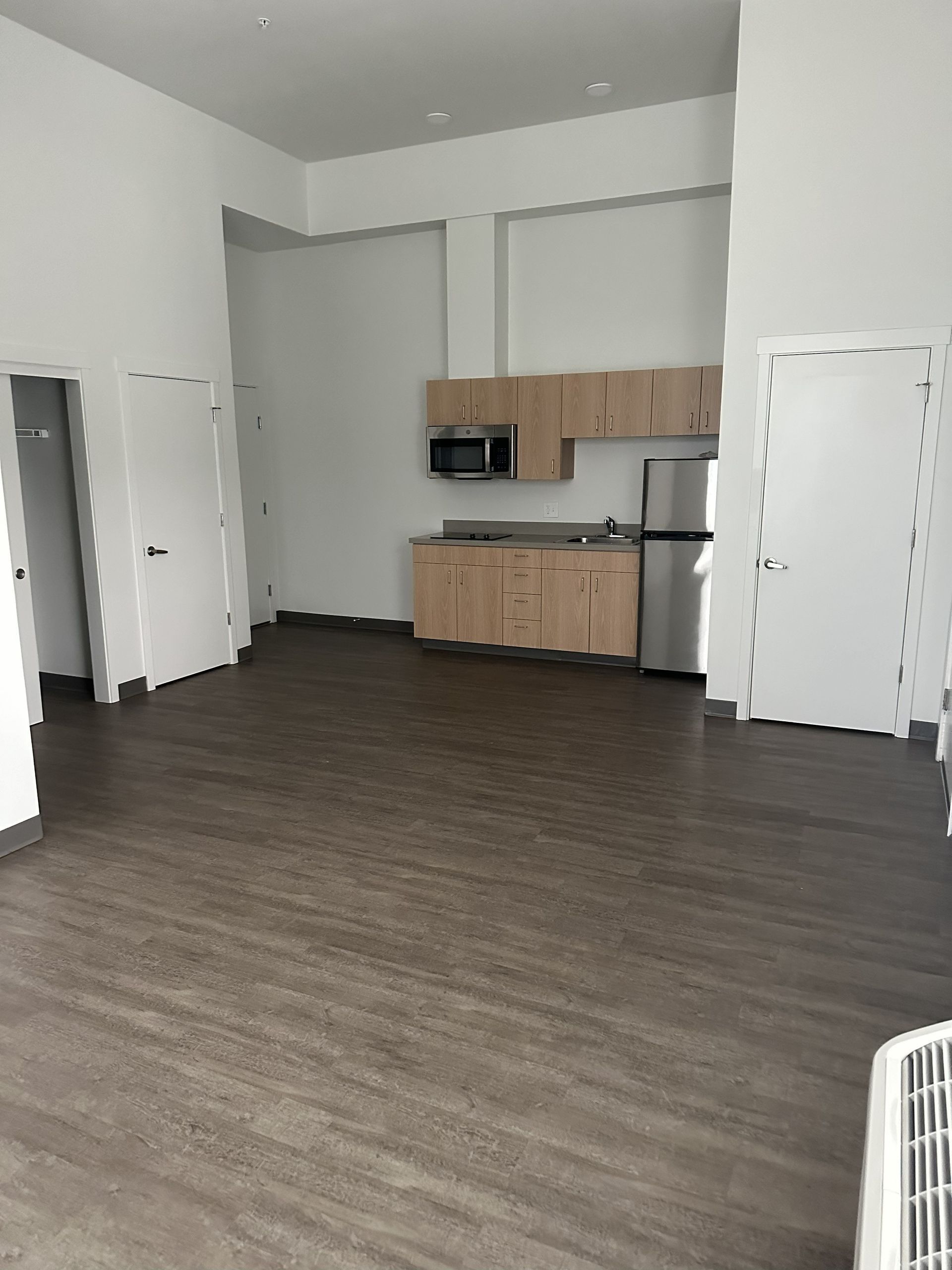 Empty room with gray wood floor, white walls, and a small kitchen alcove with cabinets and microwave.