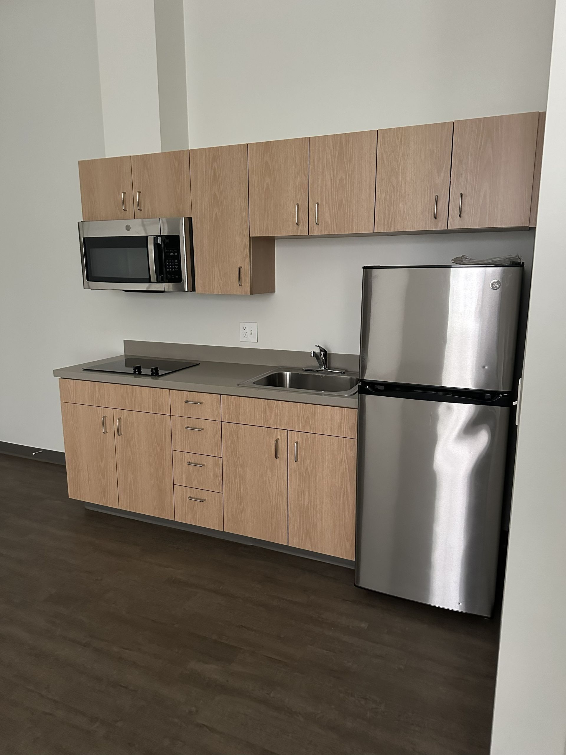 Small kitchenette with light wood cabinets, stainless steel fridge, microwave, and sink against a white wall.