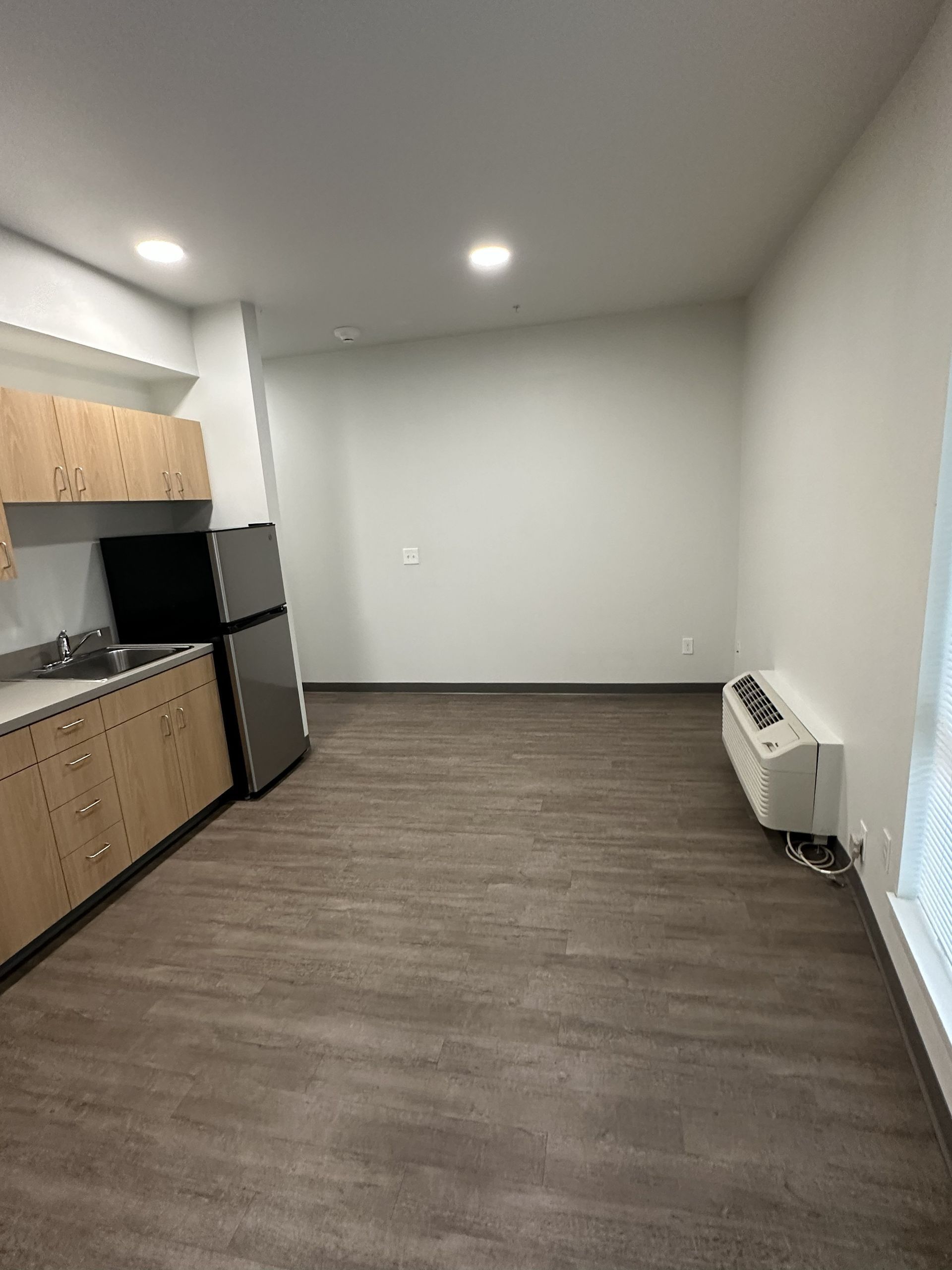 Empty modern apartment kitchen with wood cabinets, stainless fridge, and gray wood flooring.