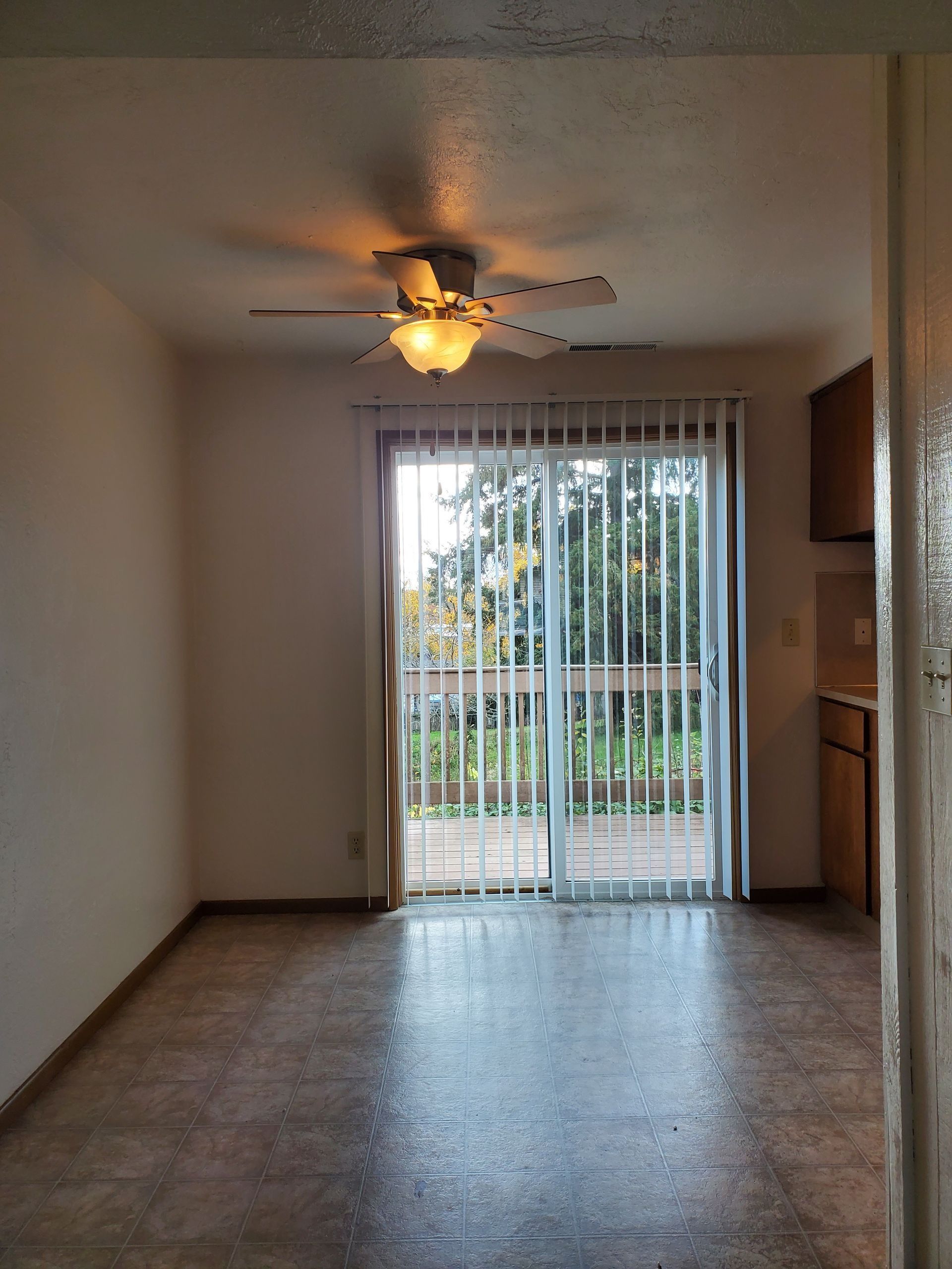 Empty living room with ceiling fan, tile floor, and sliding glass doors with vertical blinds.