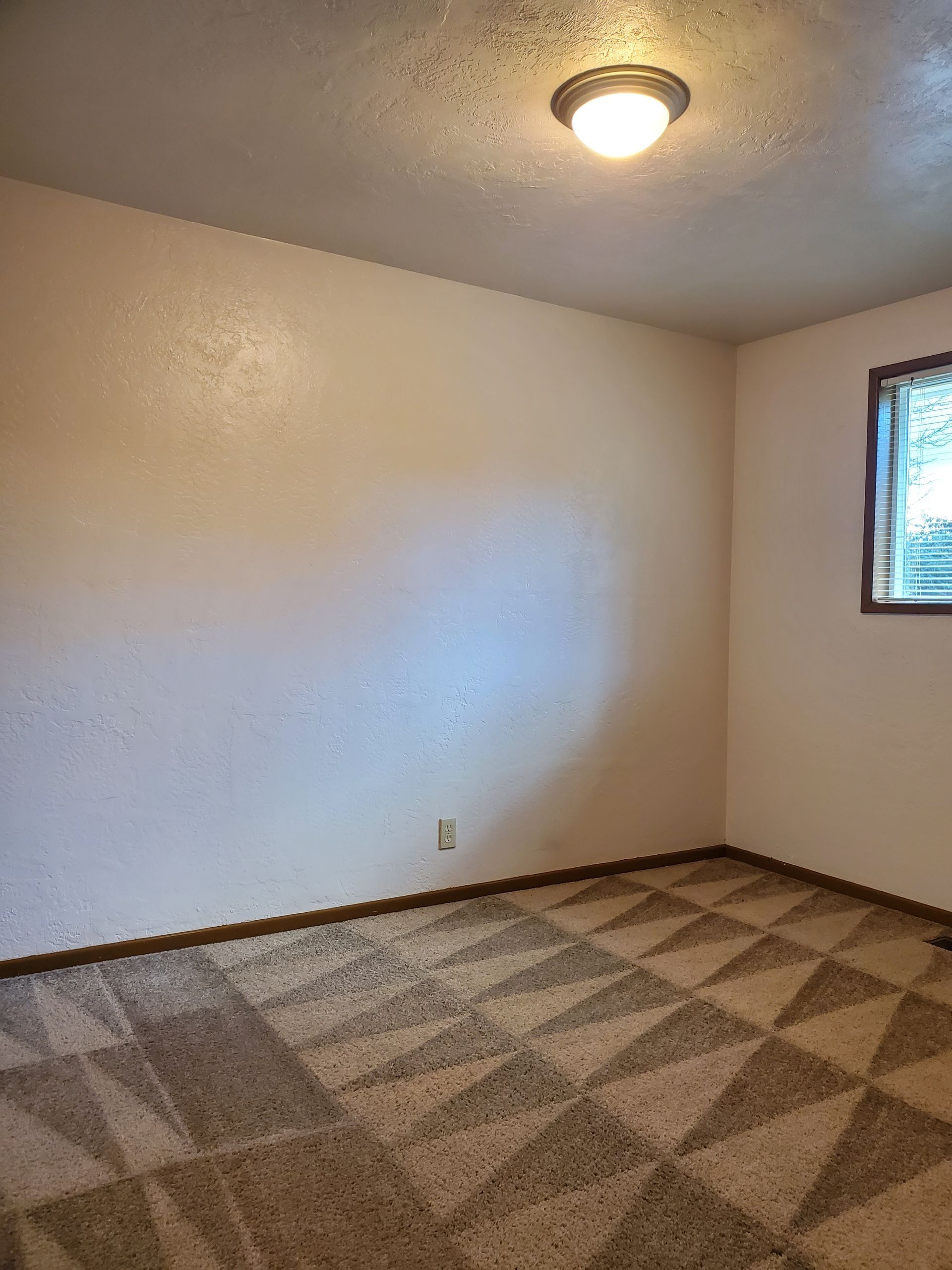 Empty beige bedroom with patterned carpet, ceiling light, and a small window.