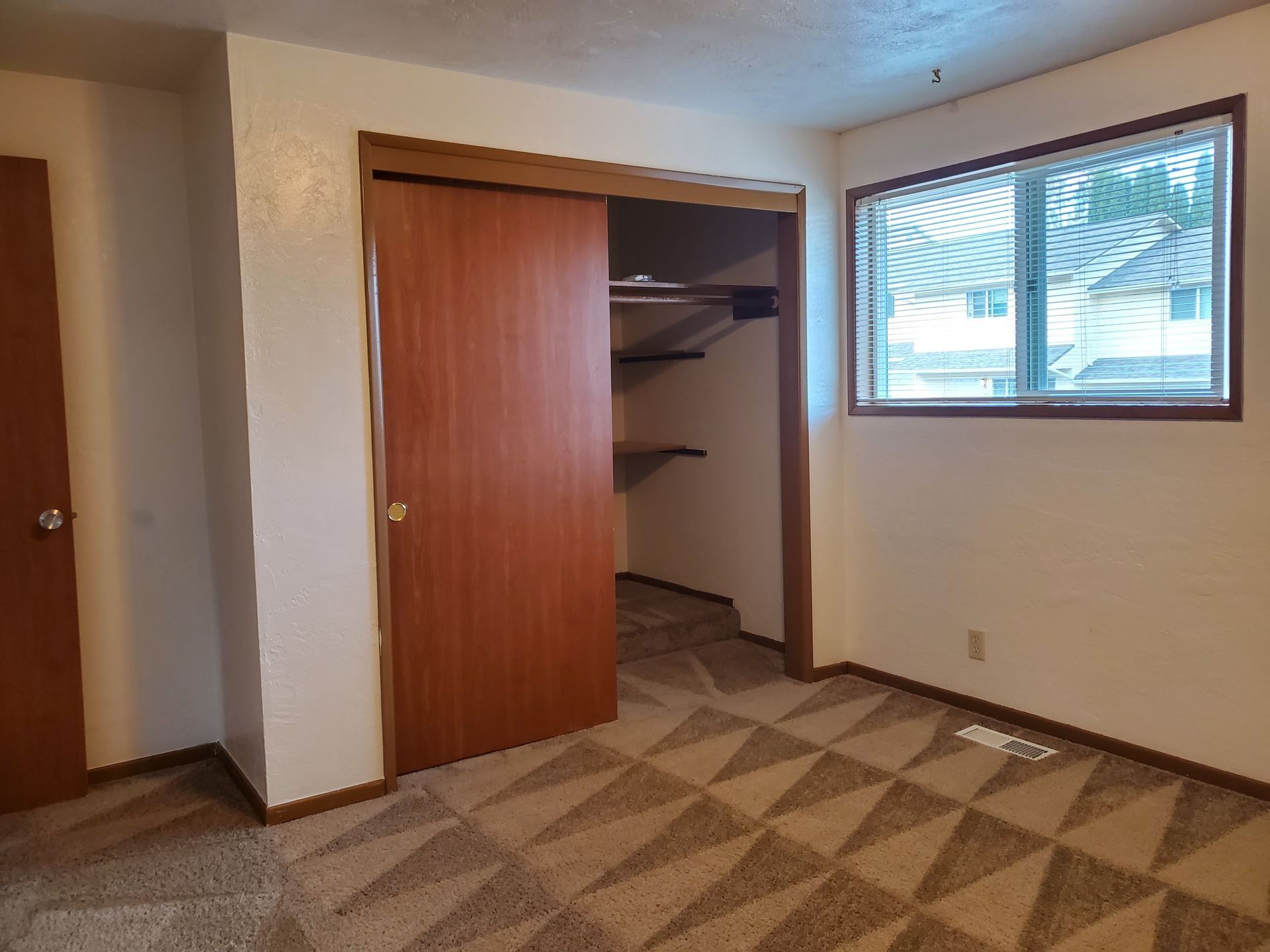 Empty beige bedroom with open closet, patterned carpet, and a window on the right