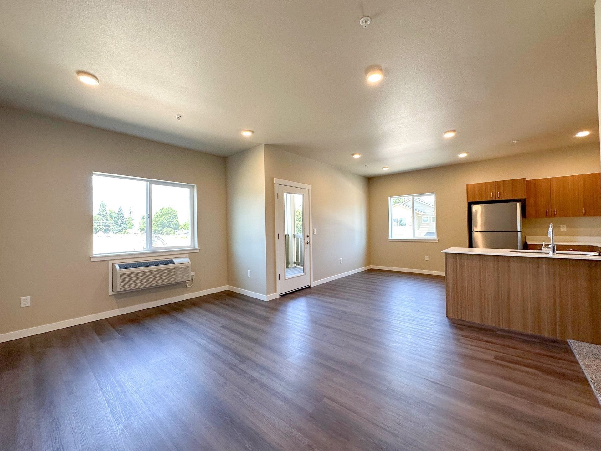 Empty modern apartment living area with wood floors, recessed lights, and kitchen on the right
