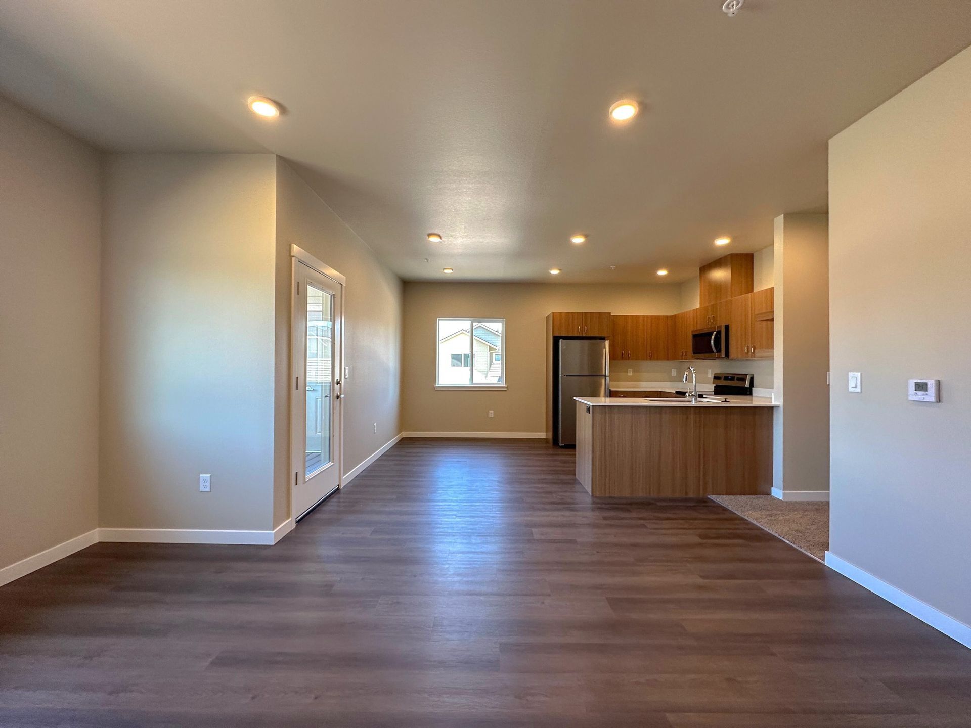 Empty open-plan living room and kitchen with wood floors, beige walls, recessed lights, and a window at the far end