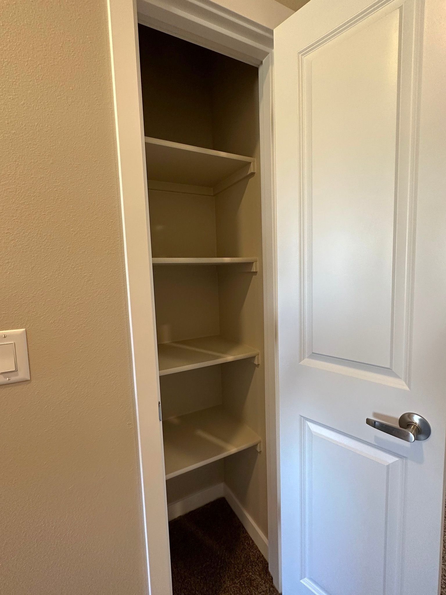Open closet with white shelves and a hanging rod, partially open white door in a beige hallway.