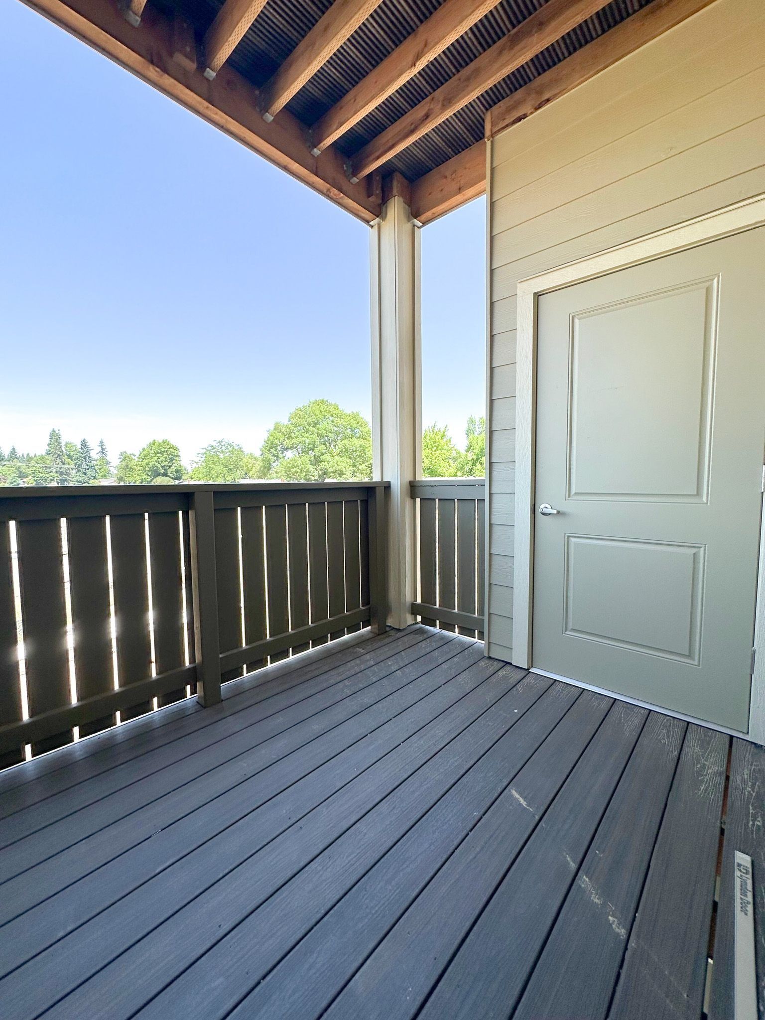 Covered wooden porch with gray floorboards, dark railing, and a white door overlooking trees.
