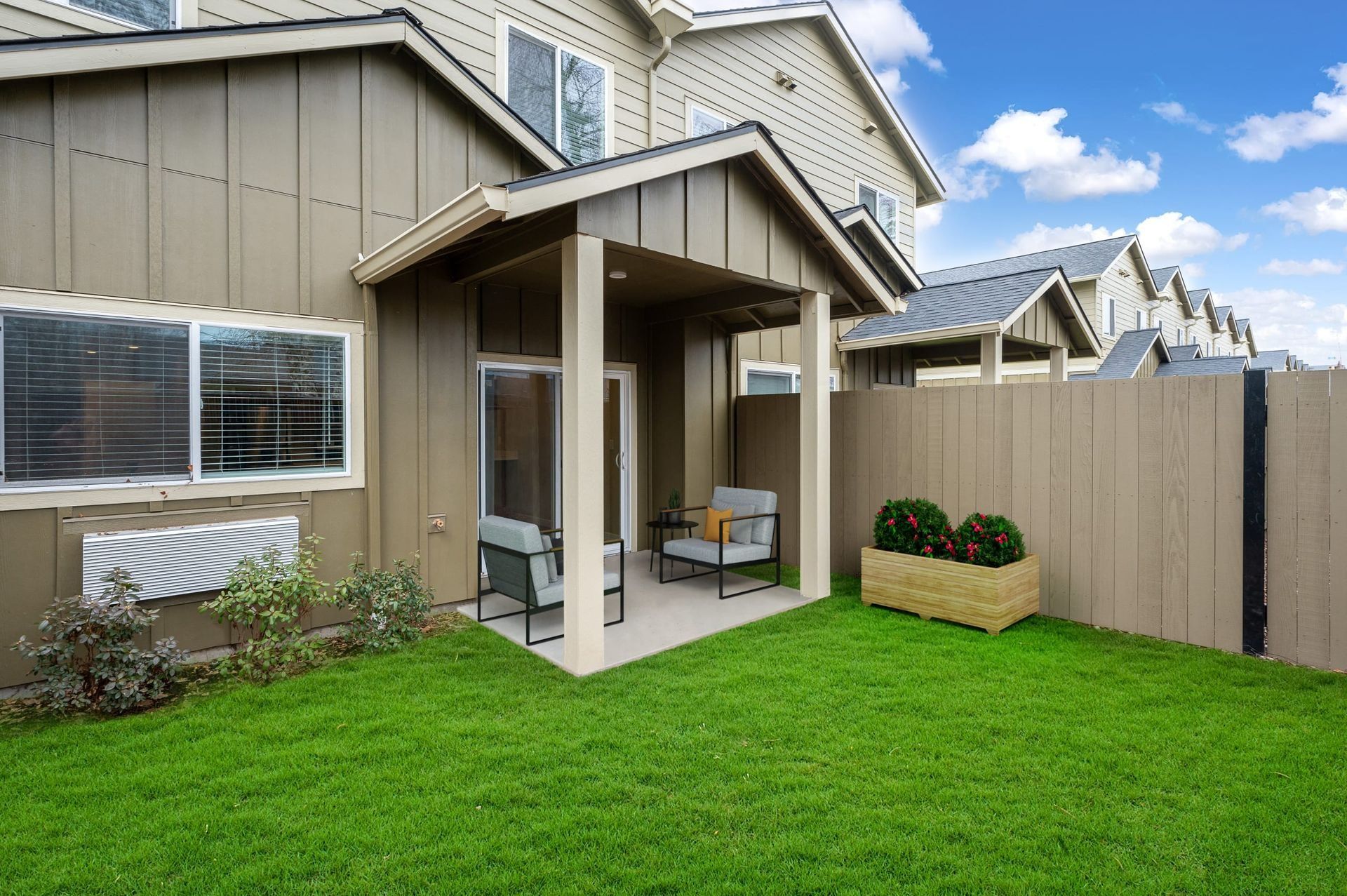 Backyard patio with chairs, green lawn, tan house, and privacy fence under a cloudy sky