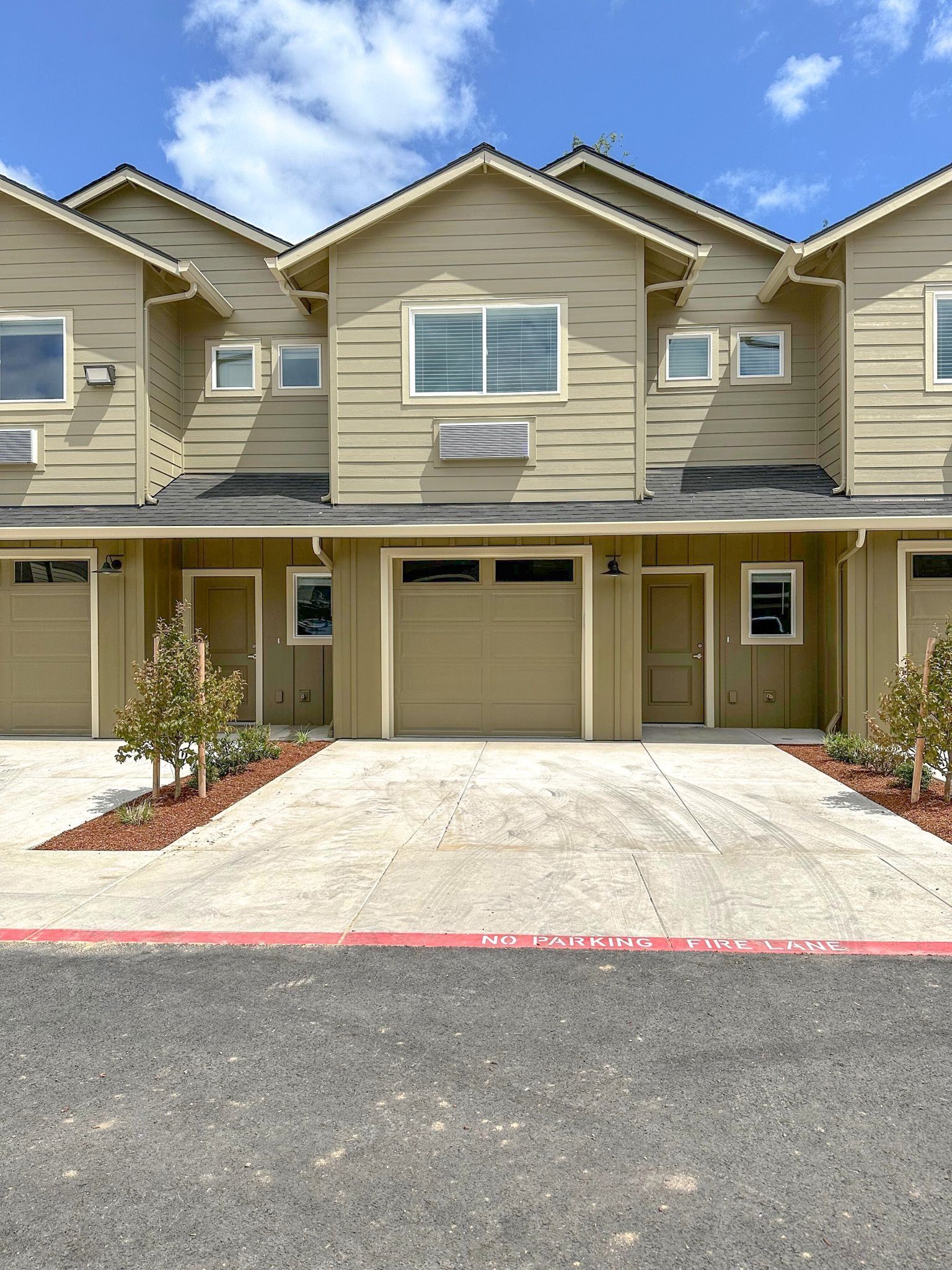 Row of beige townhomes with garages and red-mulched landscaping under a blue sky