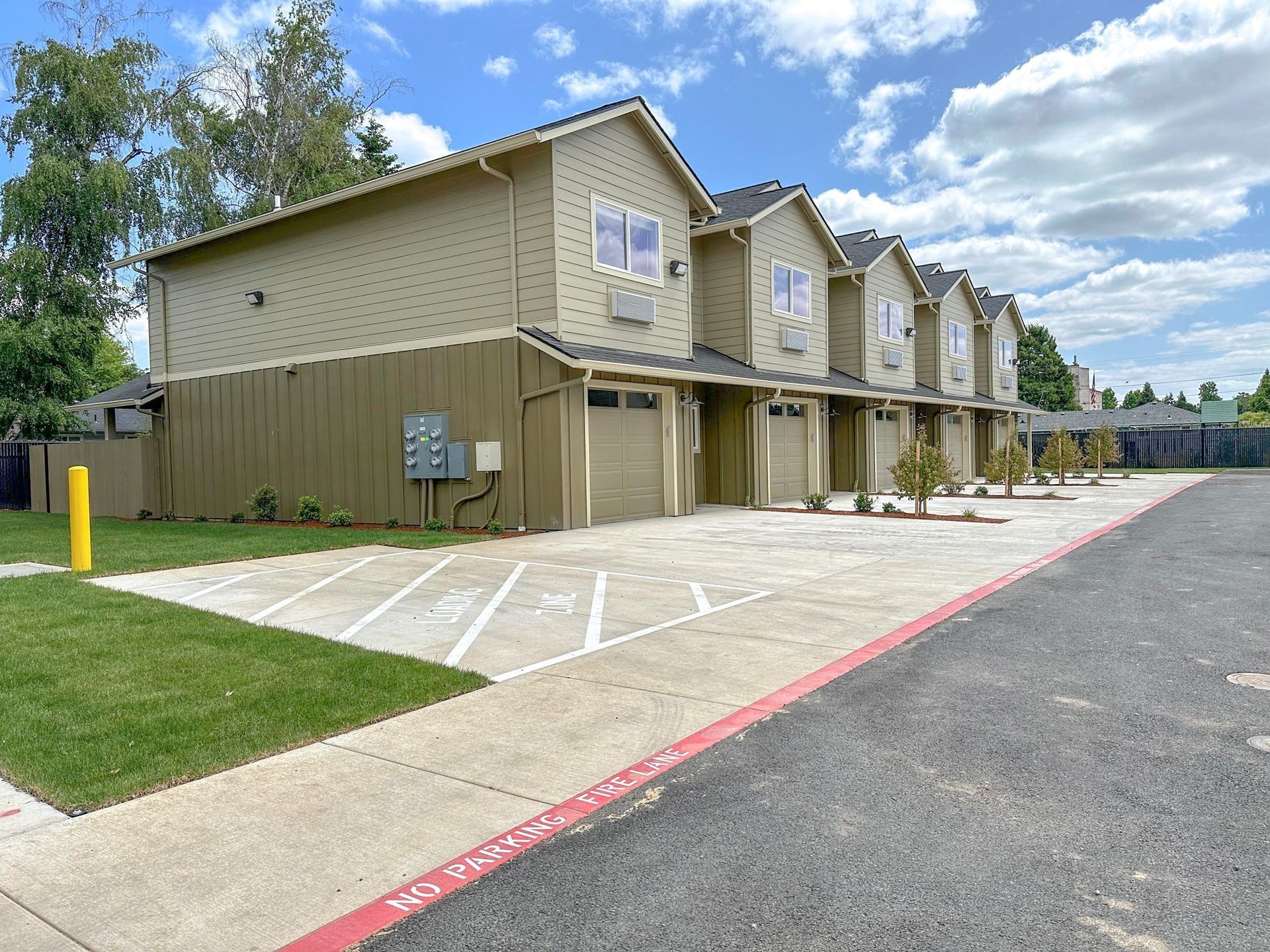 Row of beige apartment townhomes with covered parking beside a paved lot and grassy verge under a blue sky