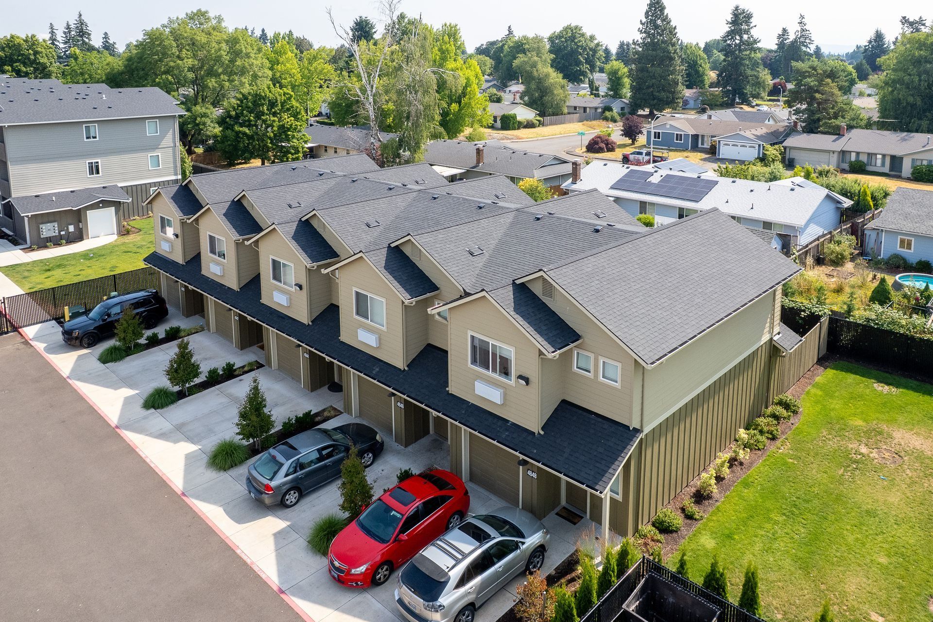 Aerial view of tan townhouses with parked cars, surrounded by trees and neighboring homes.