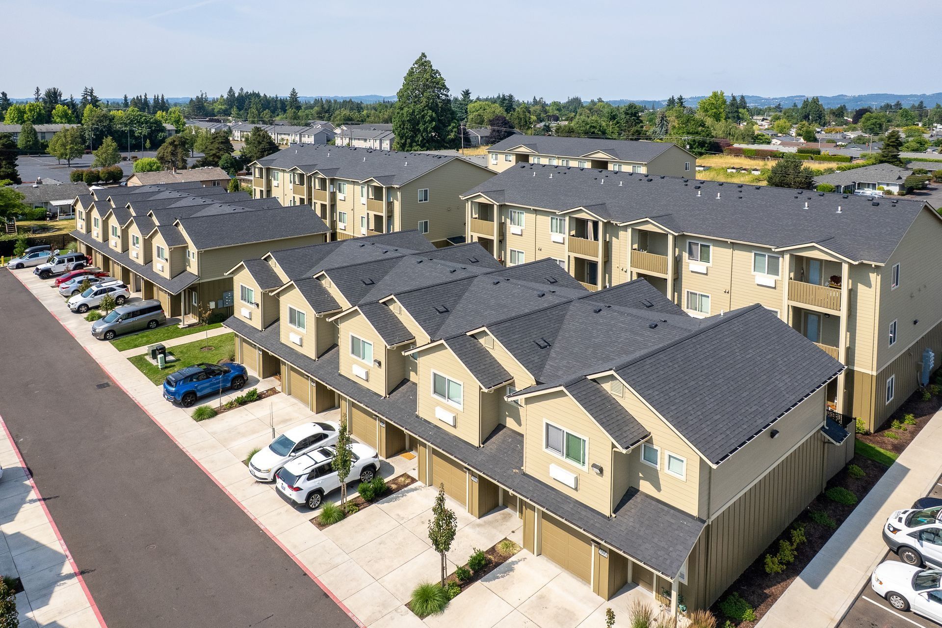 Aerial view of beige apartment buildings with dark roofs, parked cars, and tree-lined streets in a suburban complex