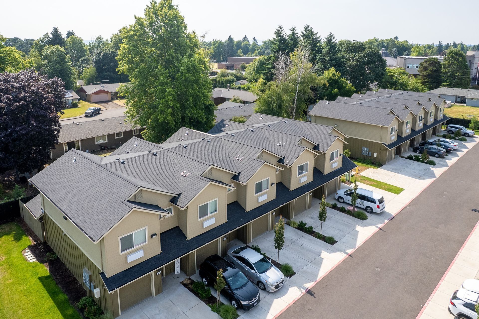 Aerial view of beige townhouses with gray roofs, parked cars, and trees along a suburban street