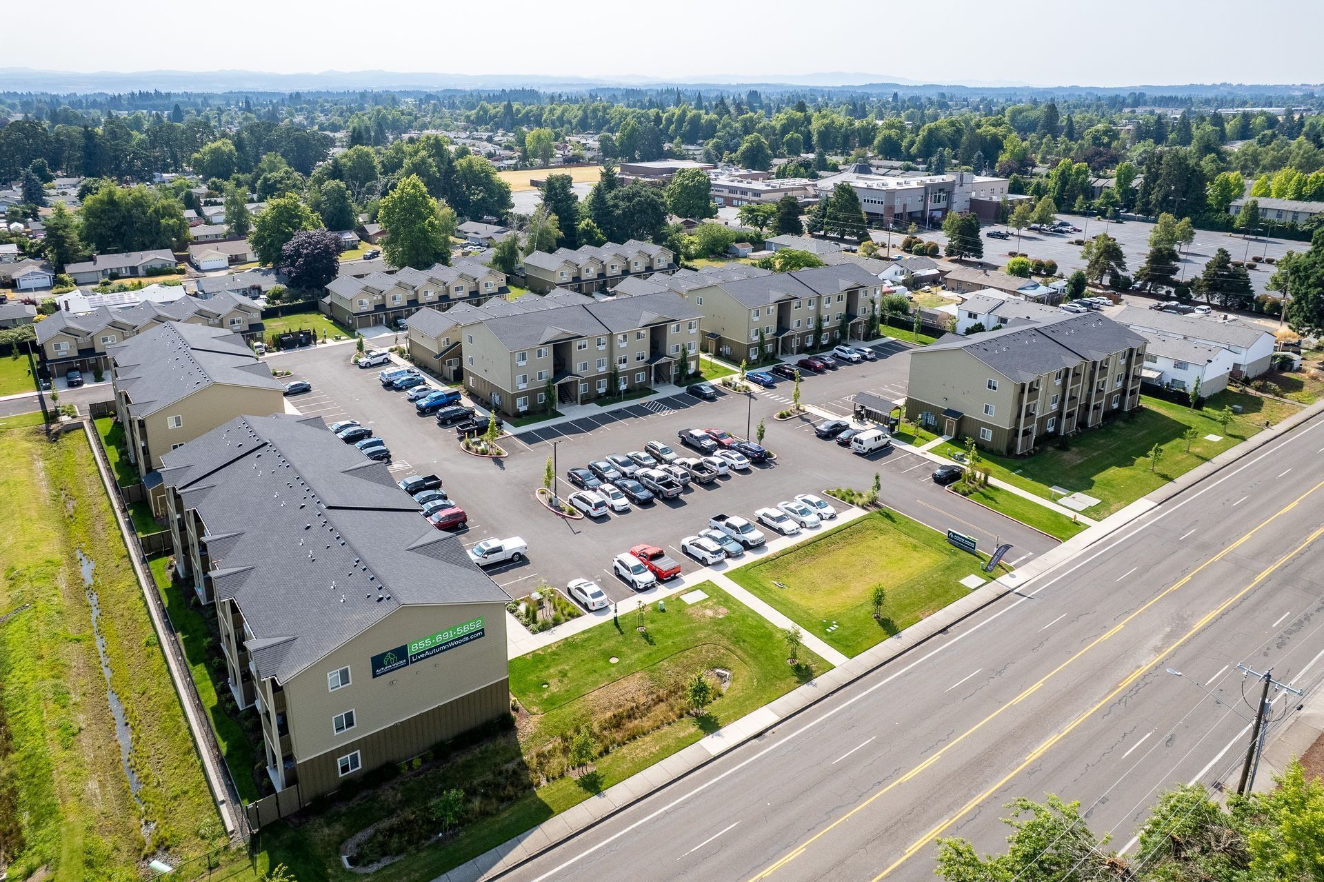 Aerial view of an apartment complex with parked cars beside a main road and trees in the distance