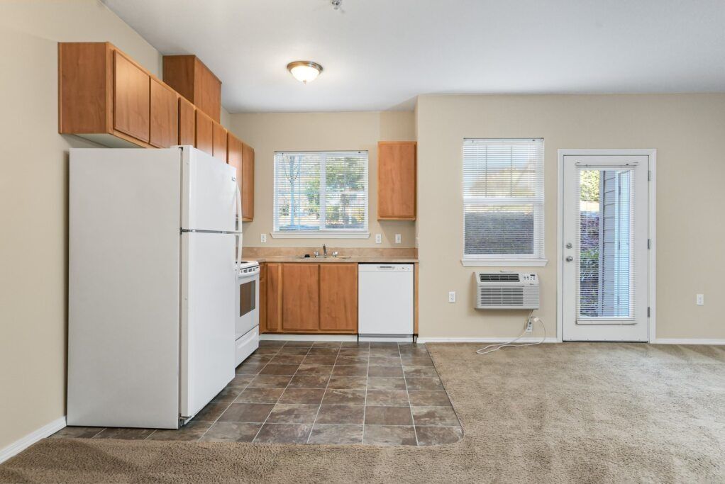Empty kitchen with white appliances, wood cabinets, tiled floor, and a door to outside.